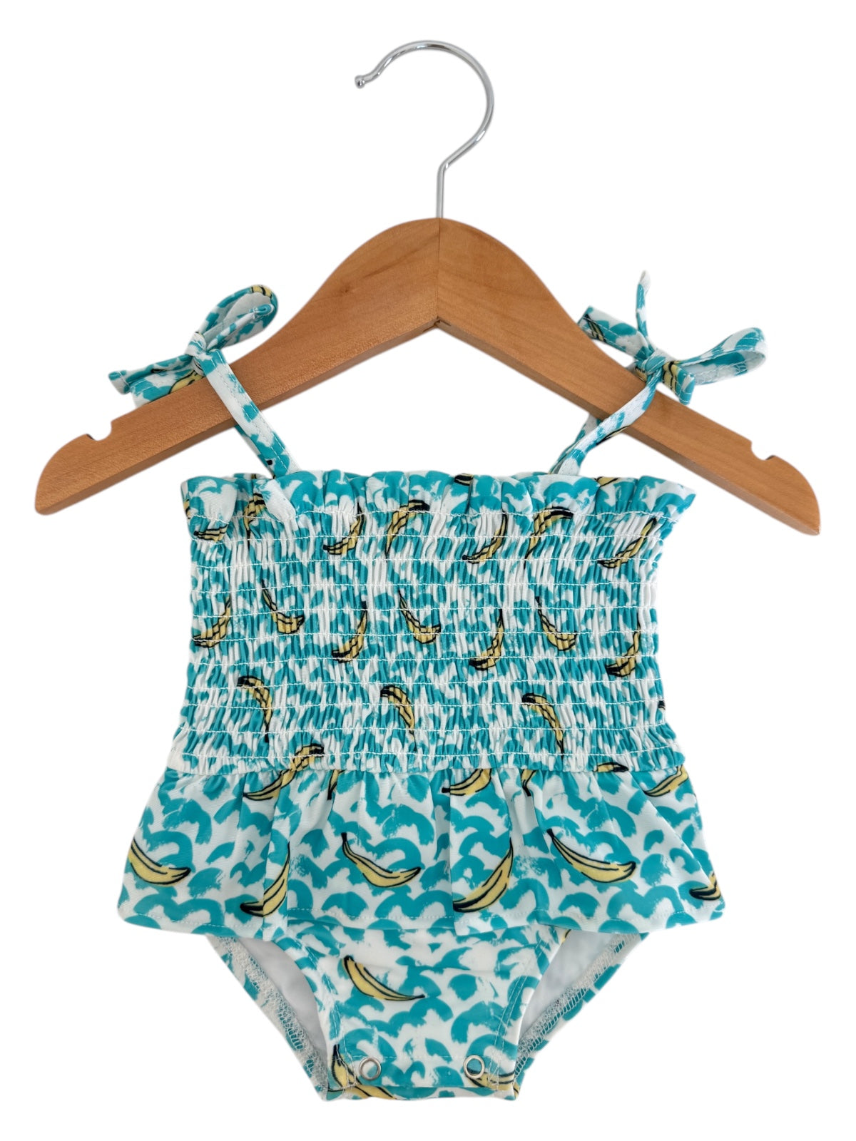 Blue swimsuit with banana print, smocked bodice, and shoulder ties, hanging on a wooden hanger.