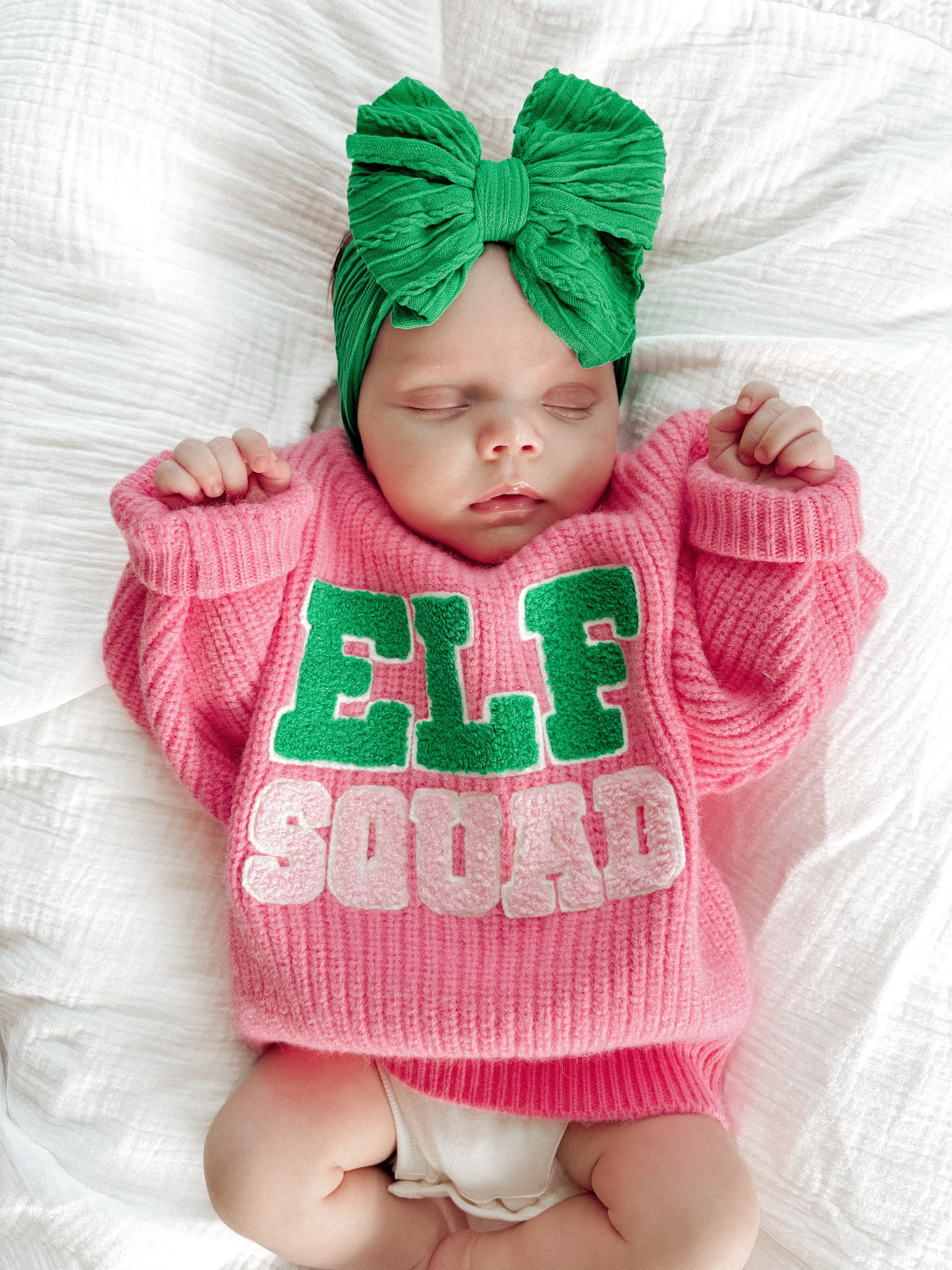 Sleeping baby in a pink "ELF SQUAD" sweater with a green bow headband, lying on a white blanket.