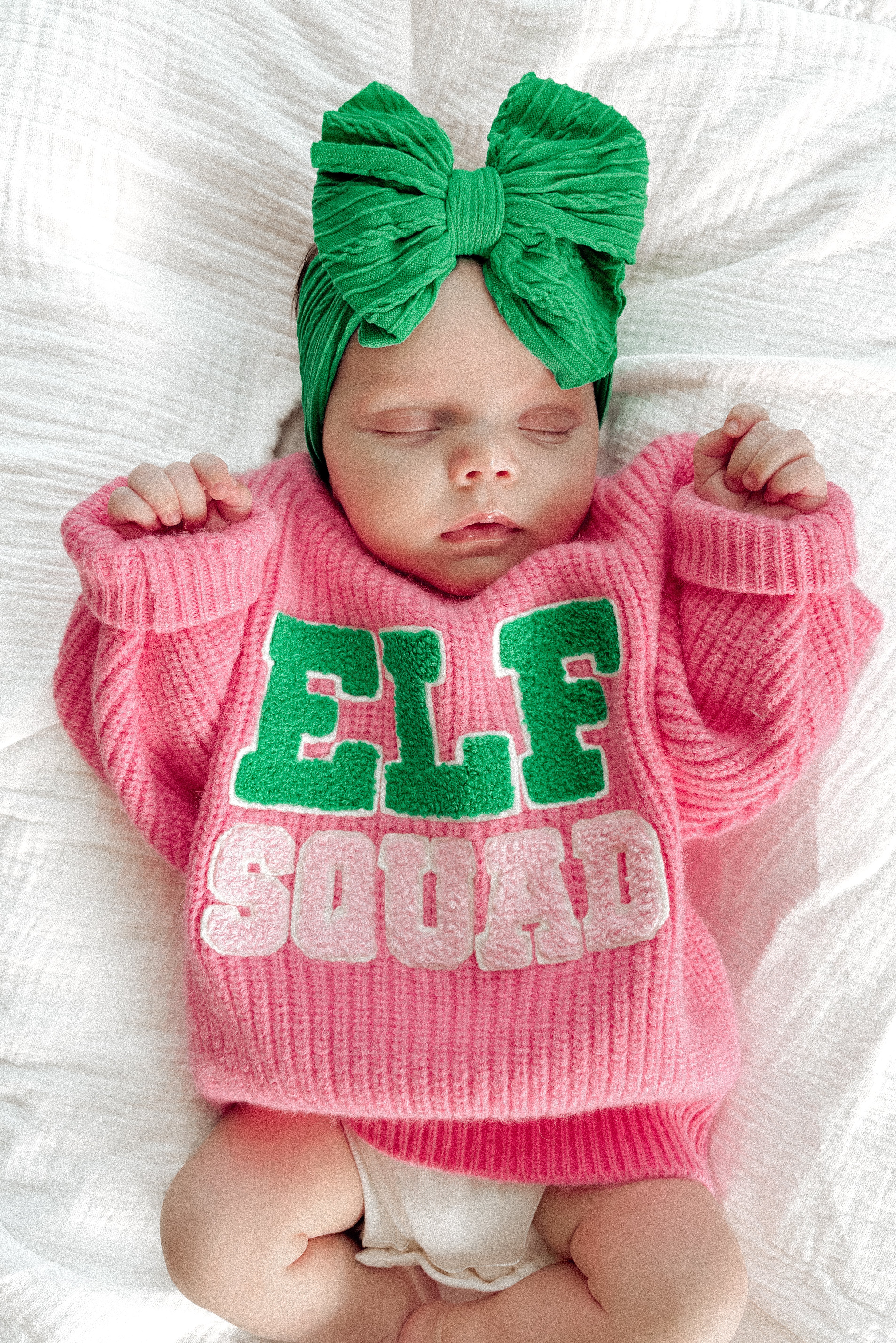 Sleeping baby in a pink "ELF SQUAD" sweater with a green bow headband, lying on a white blanket.