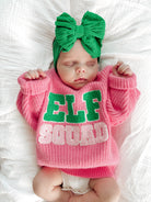 Sleeping baby in a pink "ELF SQUAD" sweater with a green bow headband, lying on a white blanket.