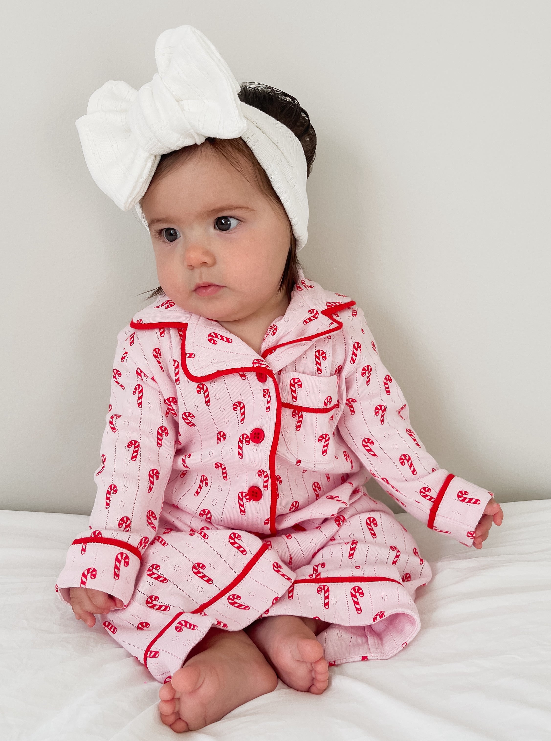 Toddler in pink candy-cane pajamas and a white headband, sitting on a white bed with a neutral background.