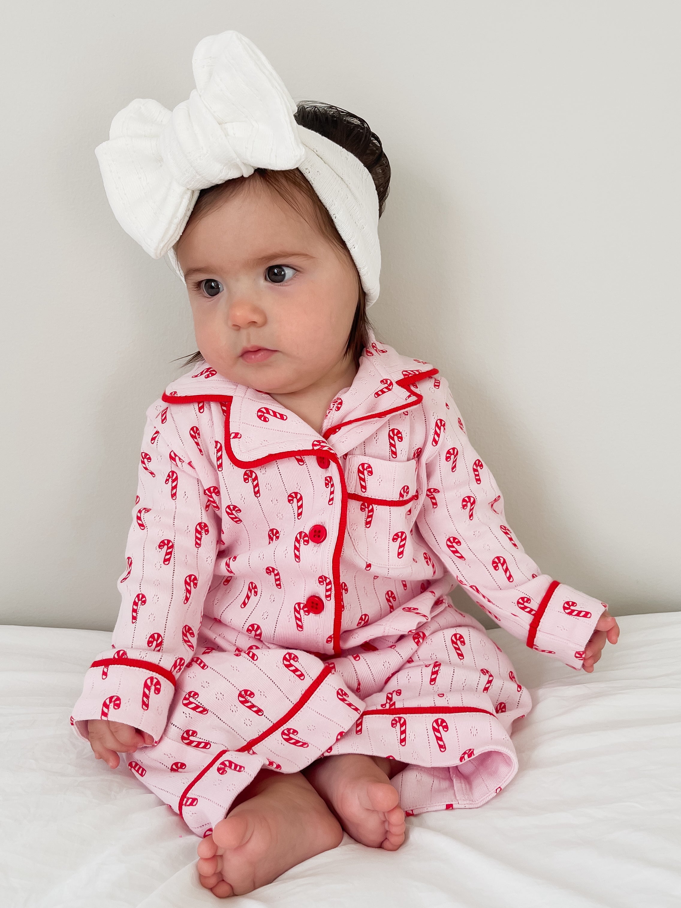 Toddler in pink candy-cane pajamas and a white headband, sitting on a white bed with a neutral background.