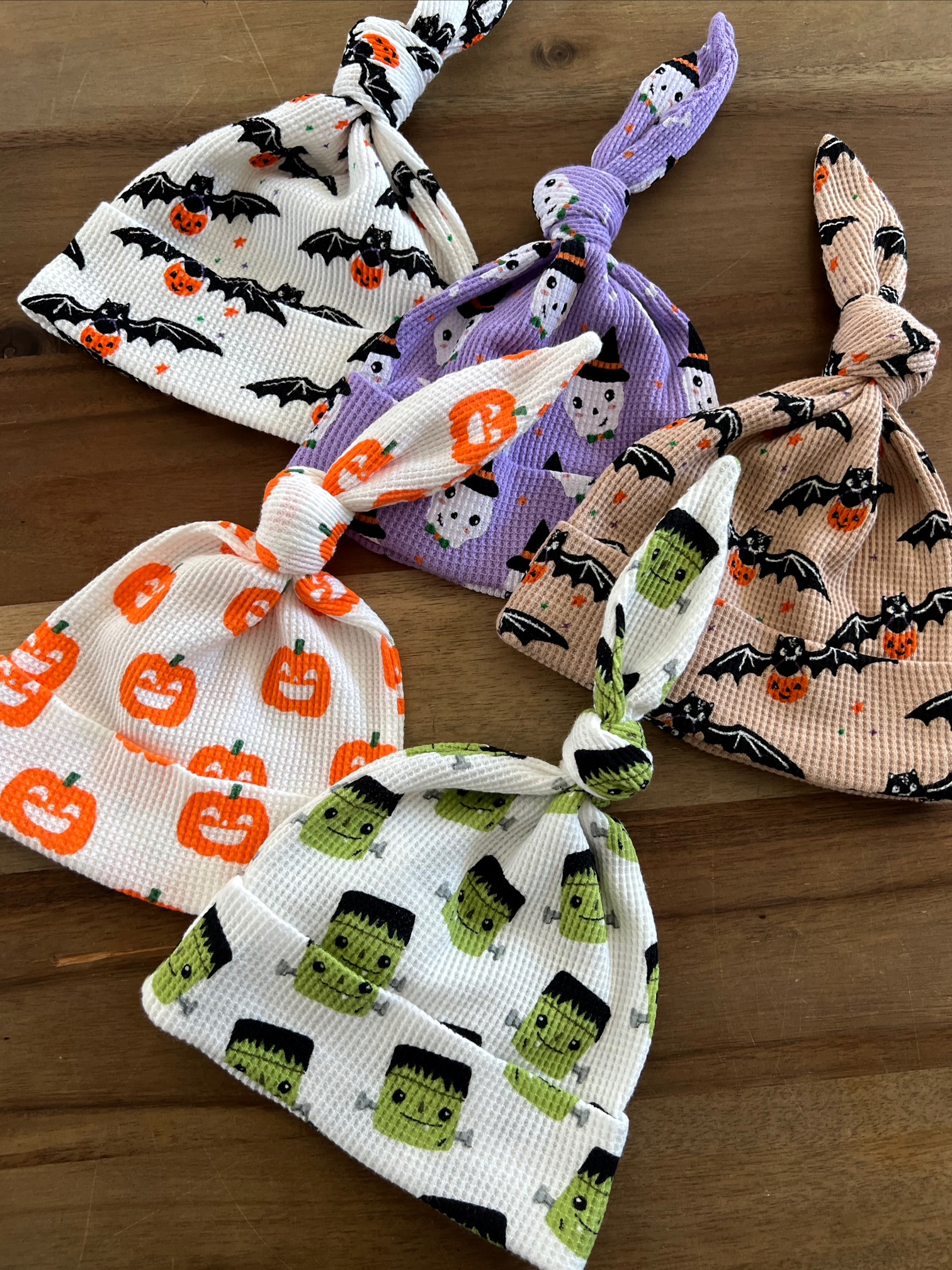 Four Halloween-themed knotted baby hats featuring bats, pumpkins, ghosts, and Frankenstein designs on a wooden surface.