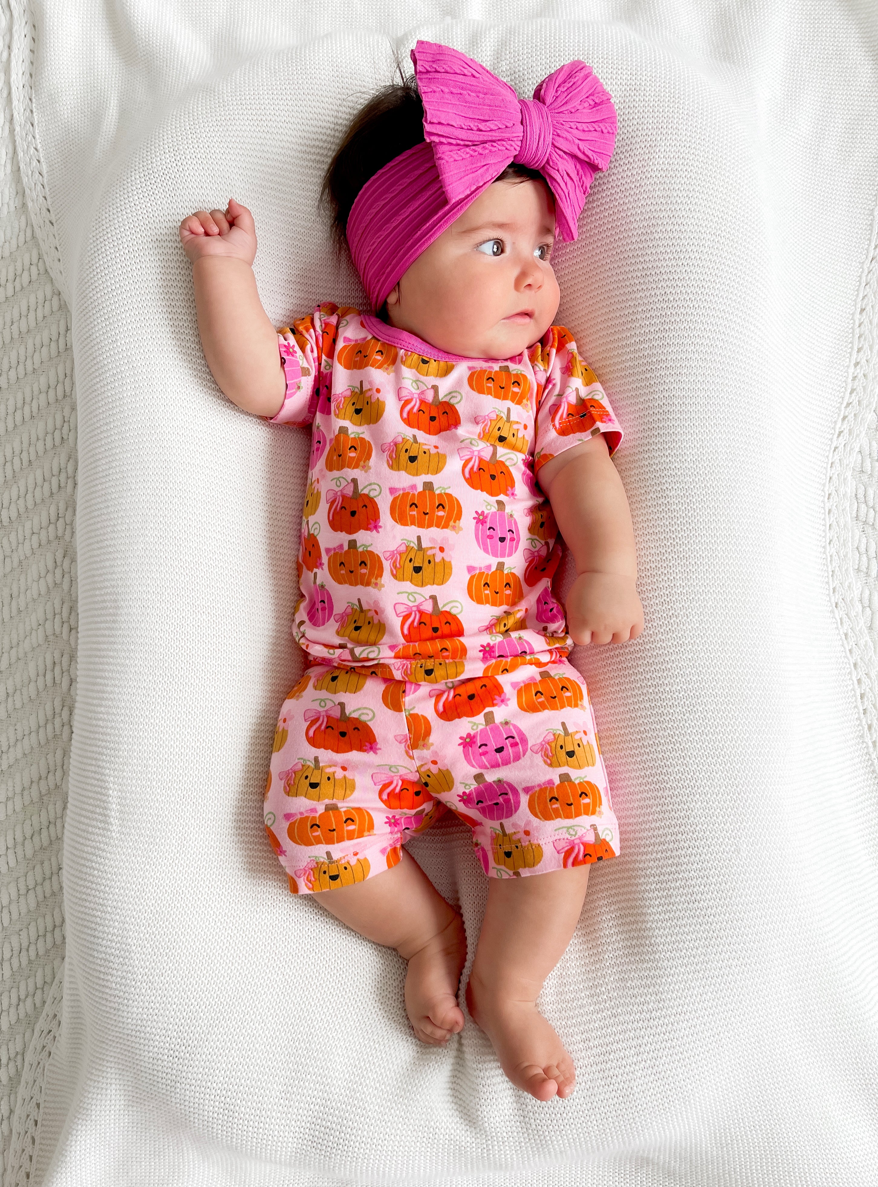 Baby girl in pumpkin-themed outfit with a pink bow headband, lying on a cozy blanket.