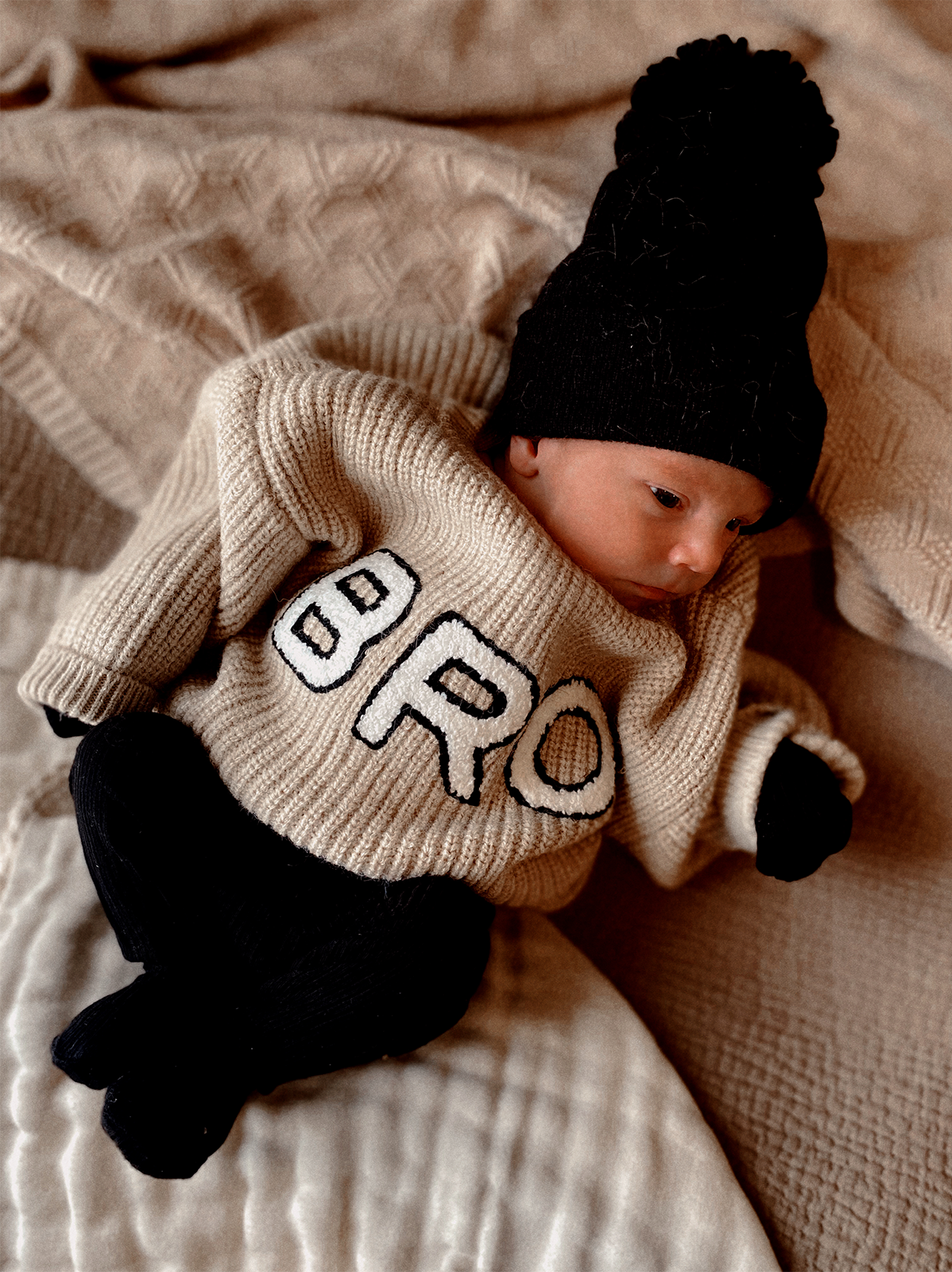 Newborn in a cozy beige sweater with "BRO" print, black pants, and a matching hat on a textured blanket.