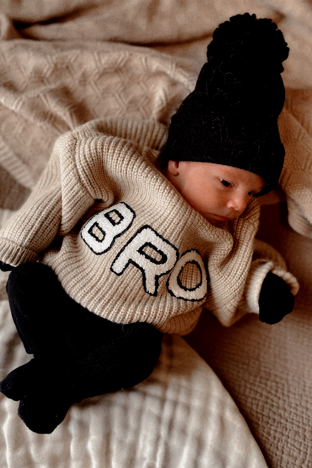 Newborn in a cozy beige sweater with "BRO" print, black pants, and a matching hat on a textured blanket.