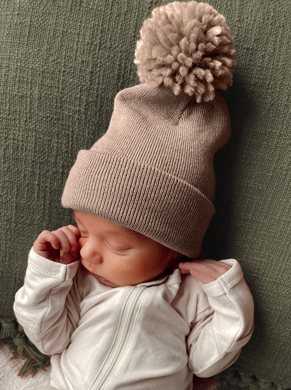 Newborn wearing a beige knit hat with a pom-pom, resting on a green textured blanket.