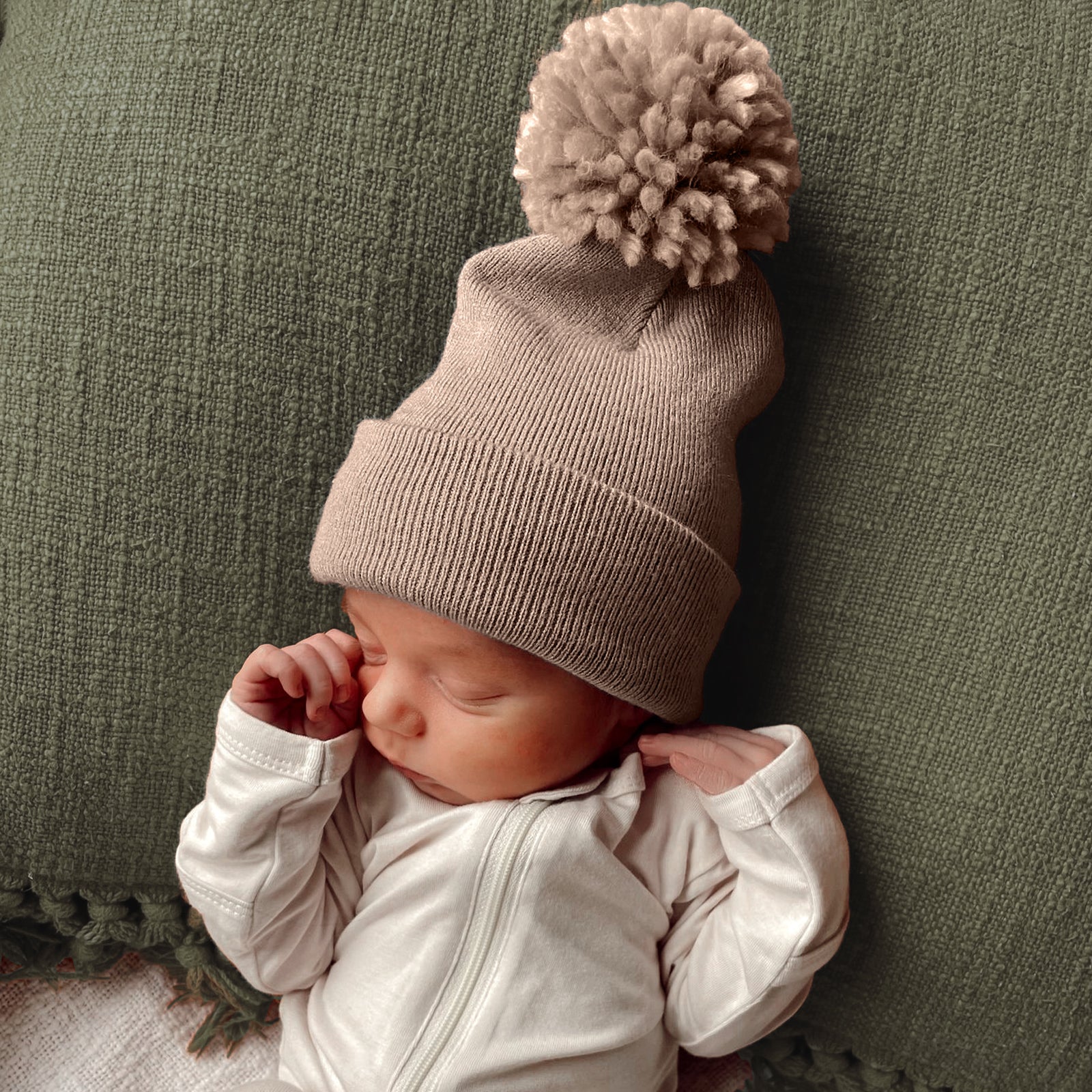 Newborn wearing a beige knit hat with a pom-pom, resting on a green textured blanket.