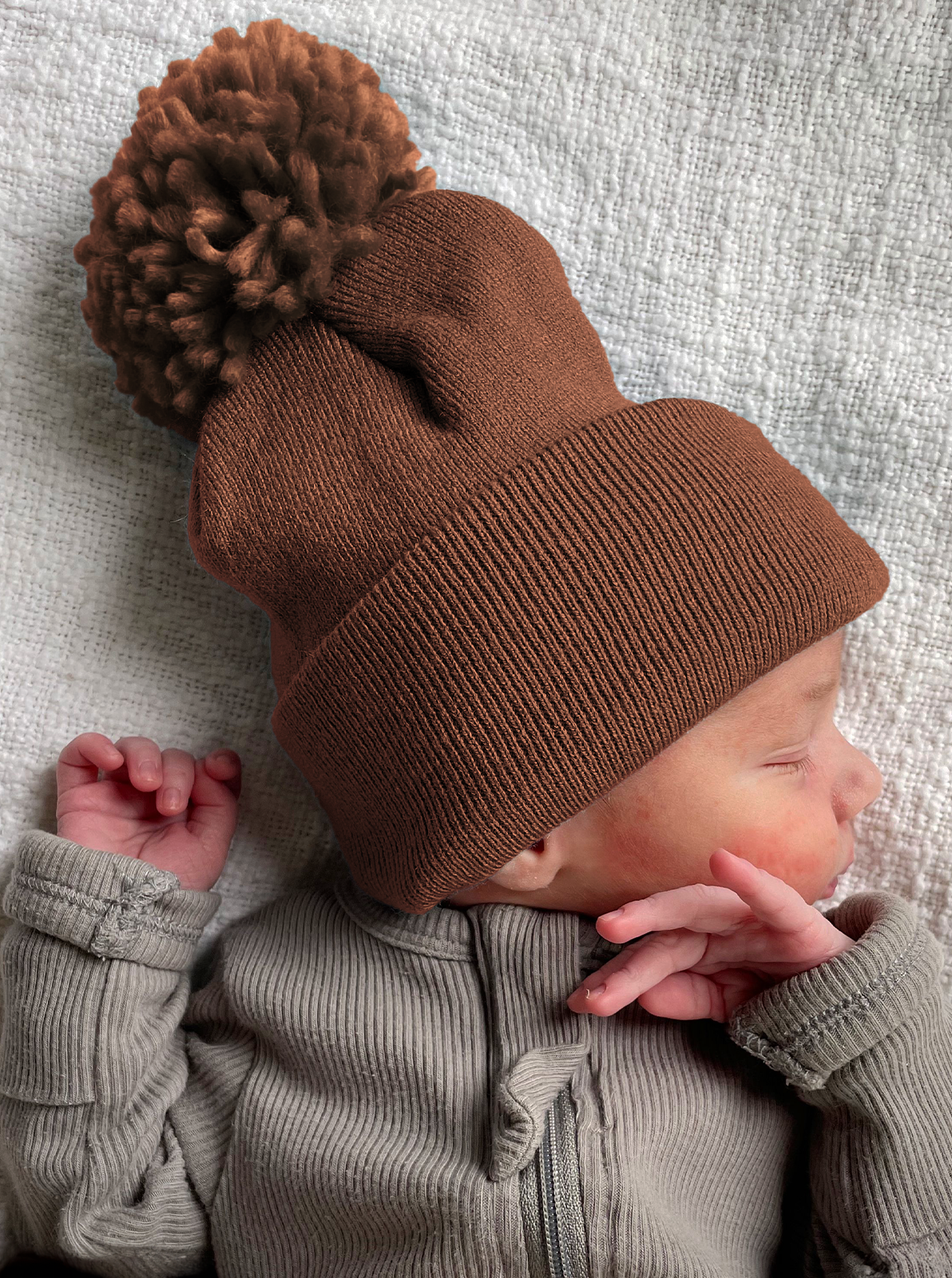 Newborn in a brown pom-pom beanie, peacefully sleeping on a textured white blanket.