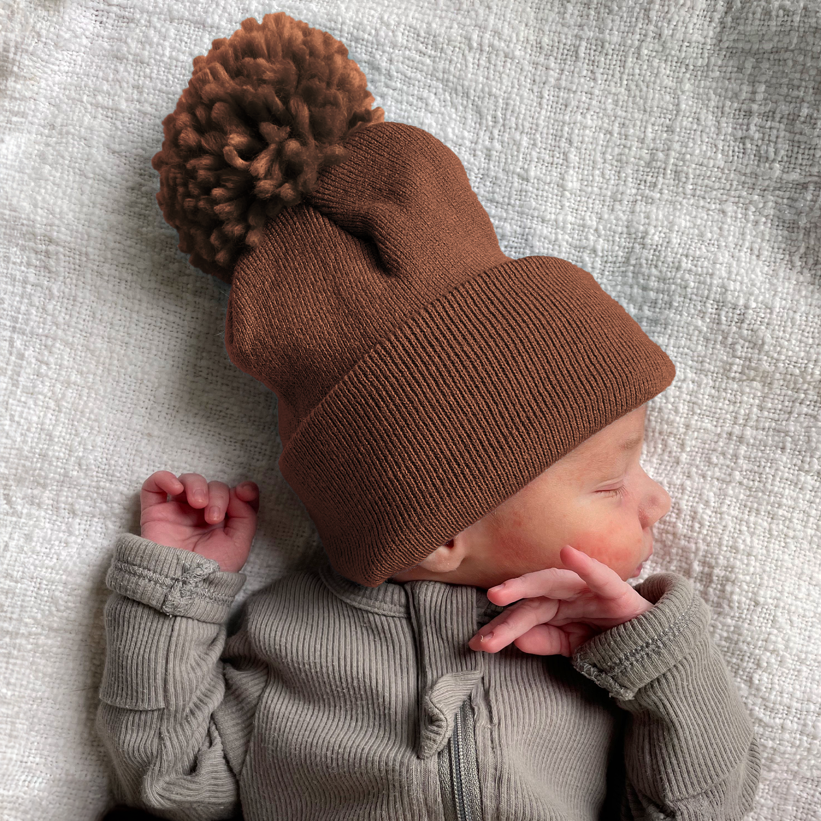 Newborn in a brown pom-pom beanie, peacefully sleeping on a textured white blanket.