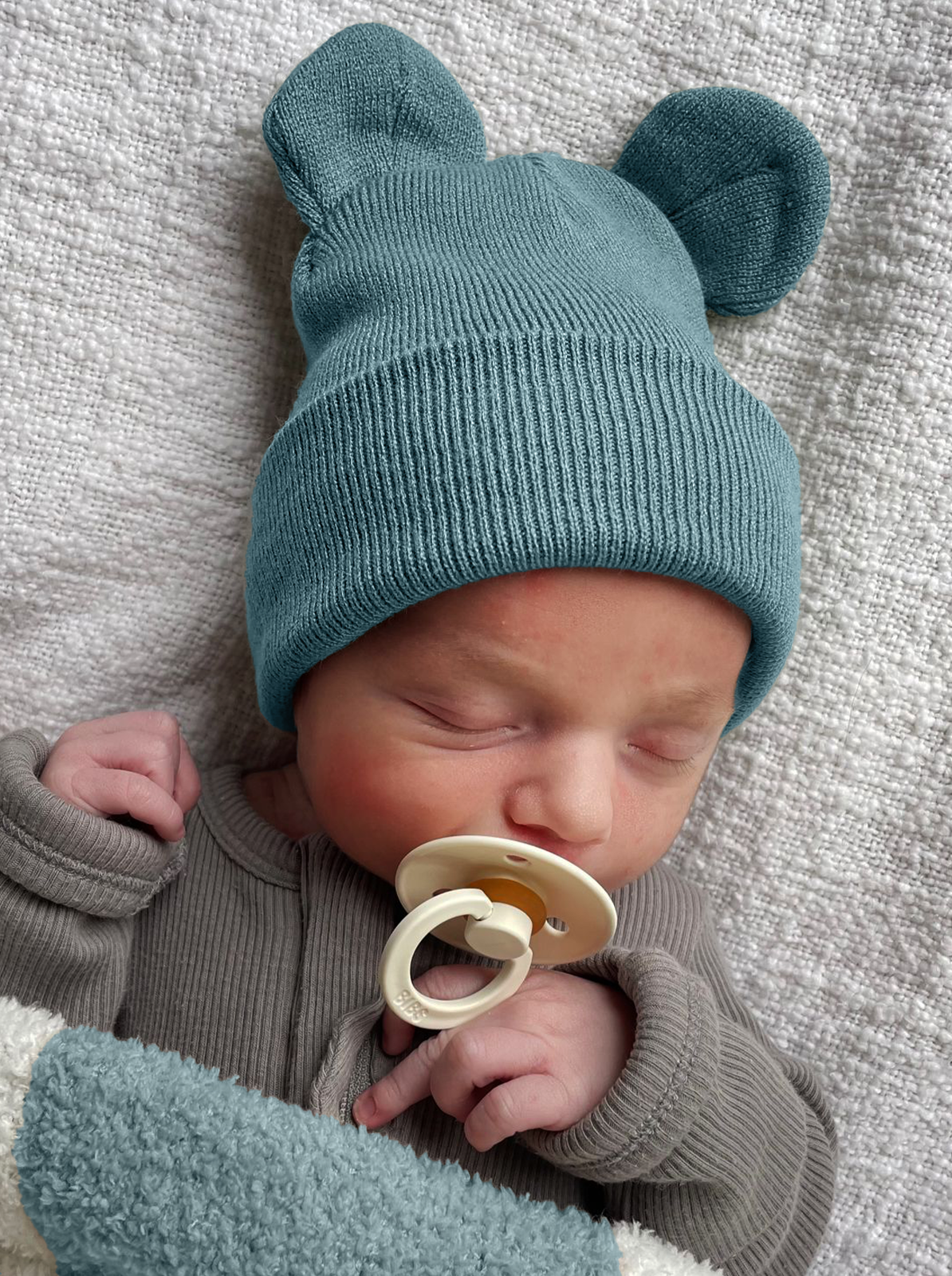 Sleeping baby with a teal bear-eared knit hat and pacifier, wrapped in a soft blanket.