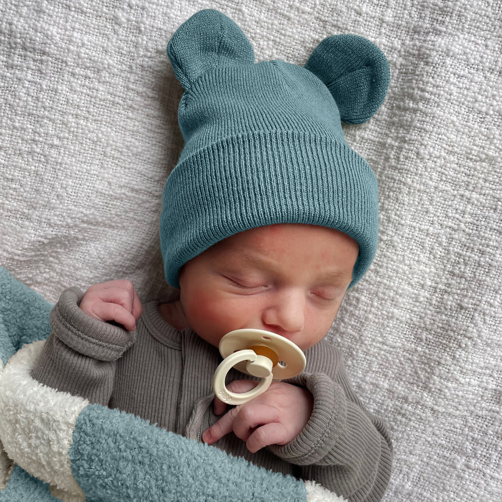 Sleeping baby with a teal bear-eared knit hat and pacifier, wrapped in a soft blanket.