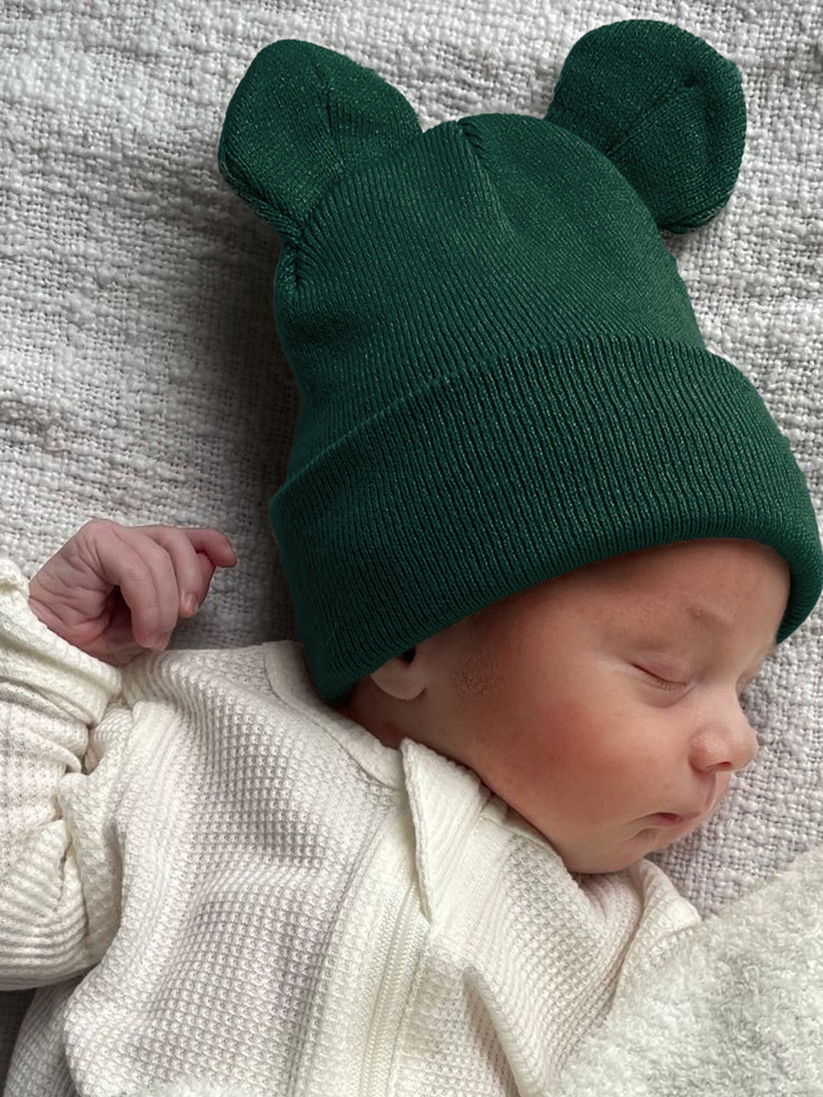 Sleeping baby in a green bear-ear beanie, wearing a white outfit, on a textured gray blanket.
