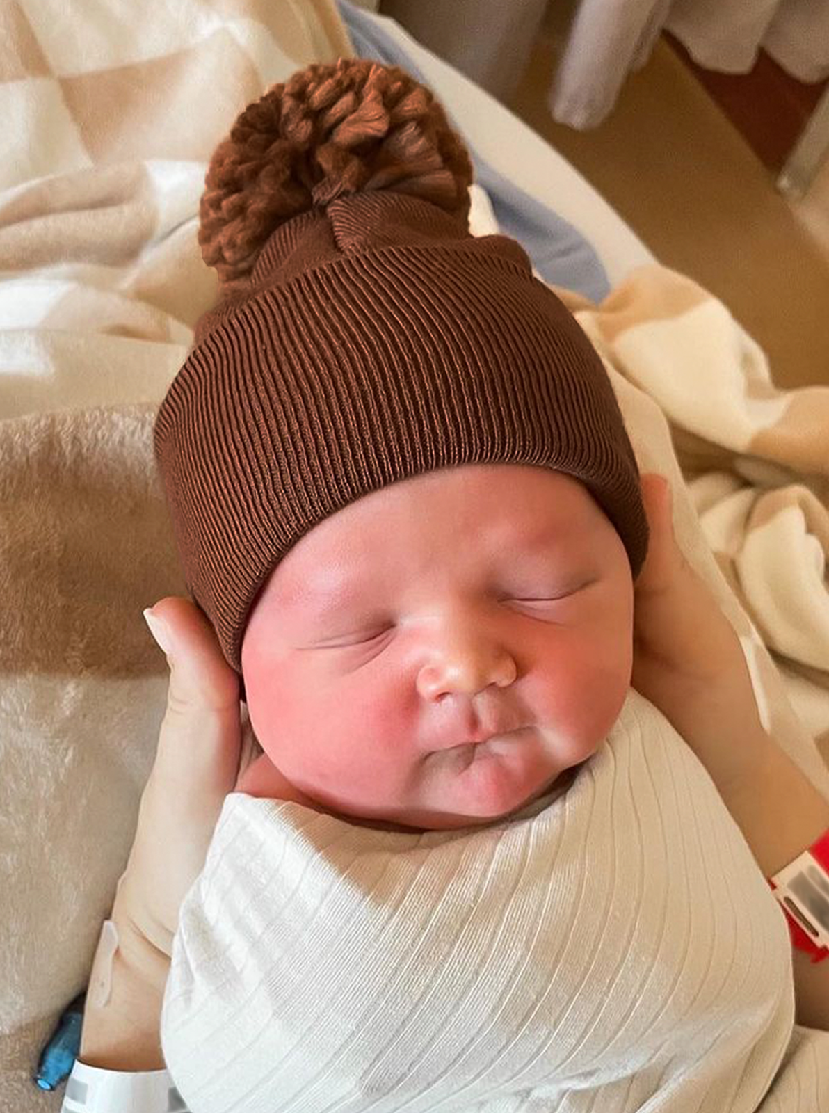 Newborn baby in a cozy brown hat, peacefully sleeping in a caregiver's hands with a soft blanket.