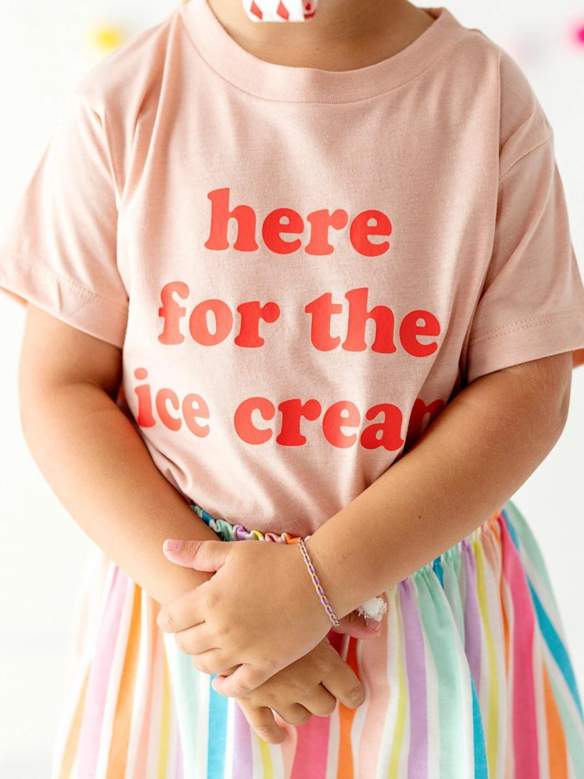 Child in a pink shirt with red text reading "here for the ice cream," wearing a colorful striped skirt. Hands crossed.