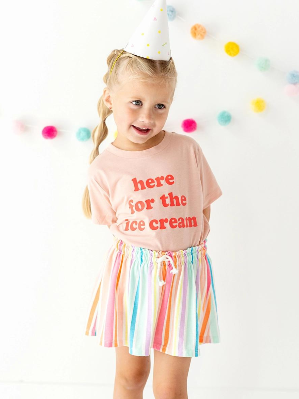 Girl wearing a party hat, pink shirt saying "here for the ice cream," and colorful striped skirt, smiling in a bright setting.
