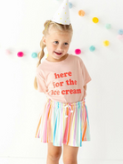 Girl wearing a party hat, pink shirt saying "here for the ice cream," and colorful striped skirt, smiling in a bright setting.