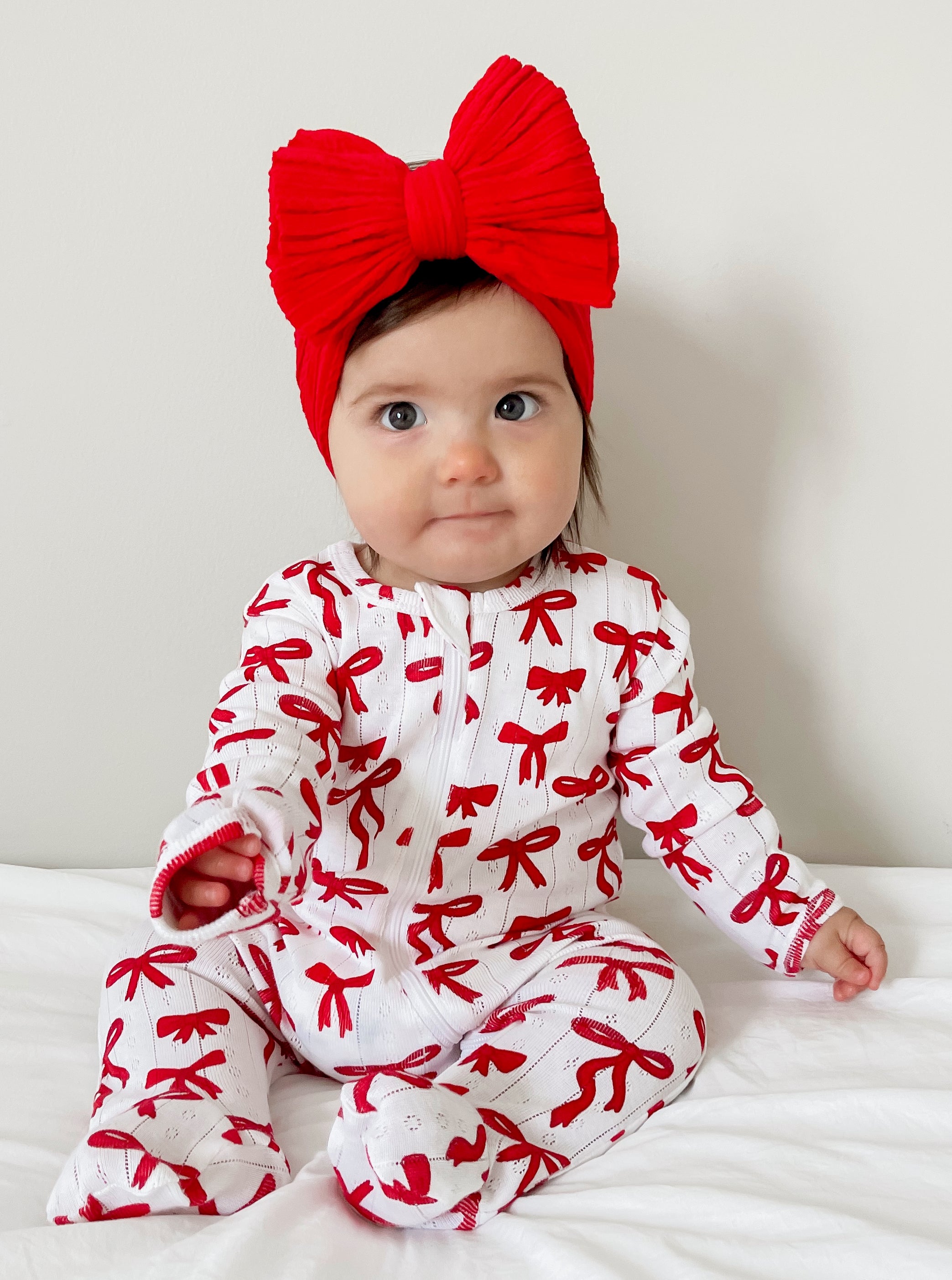 Smiling baby wearing a red bow headband and a white outfit with red bows, sitting on a white blanket.