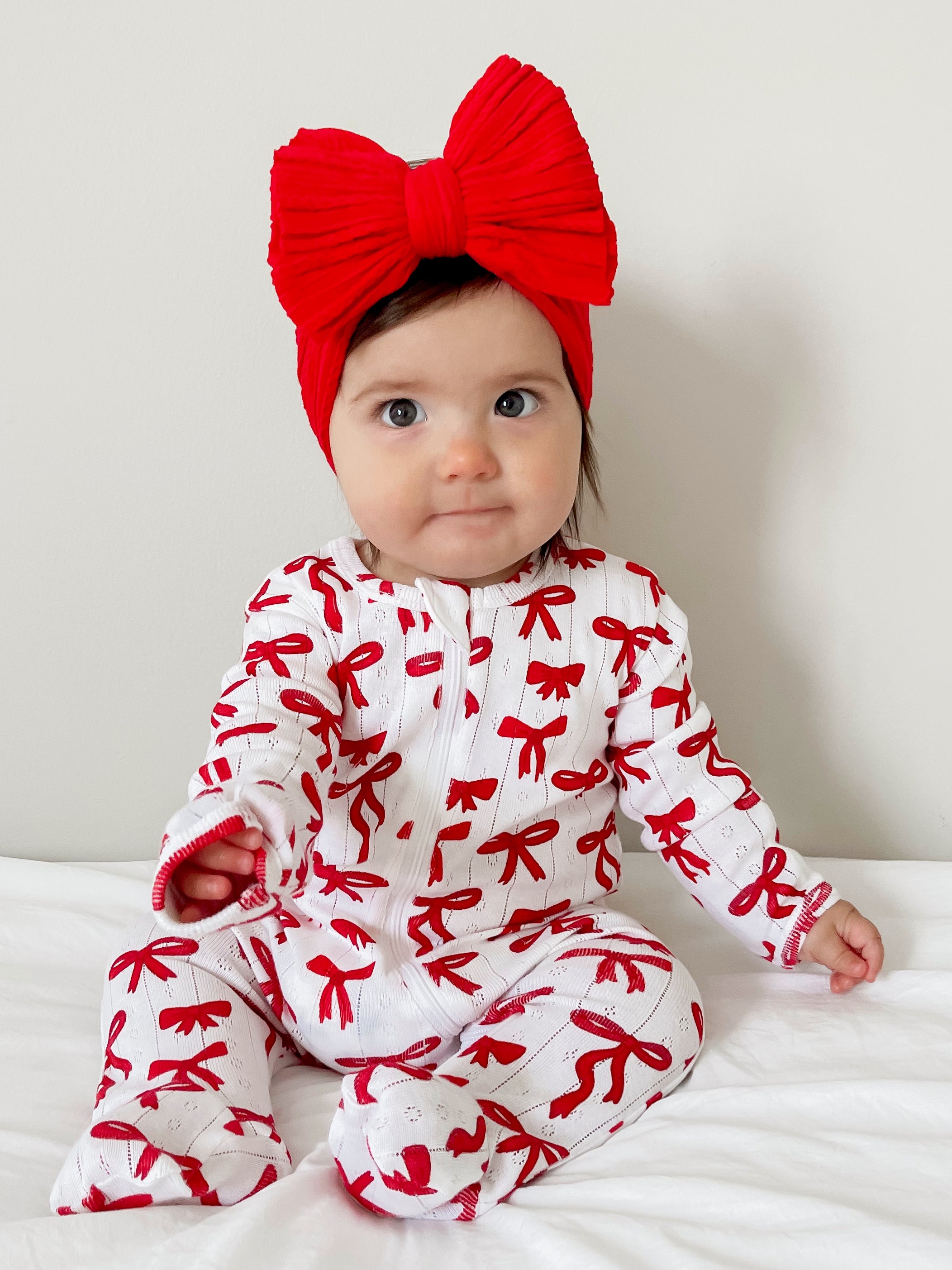 Smiling baby wearing a red bow headband and a white outfit with red bows, sitting on a white blanket.