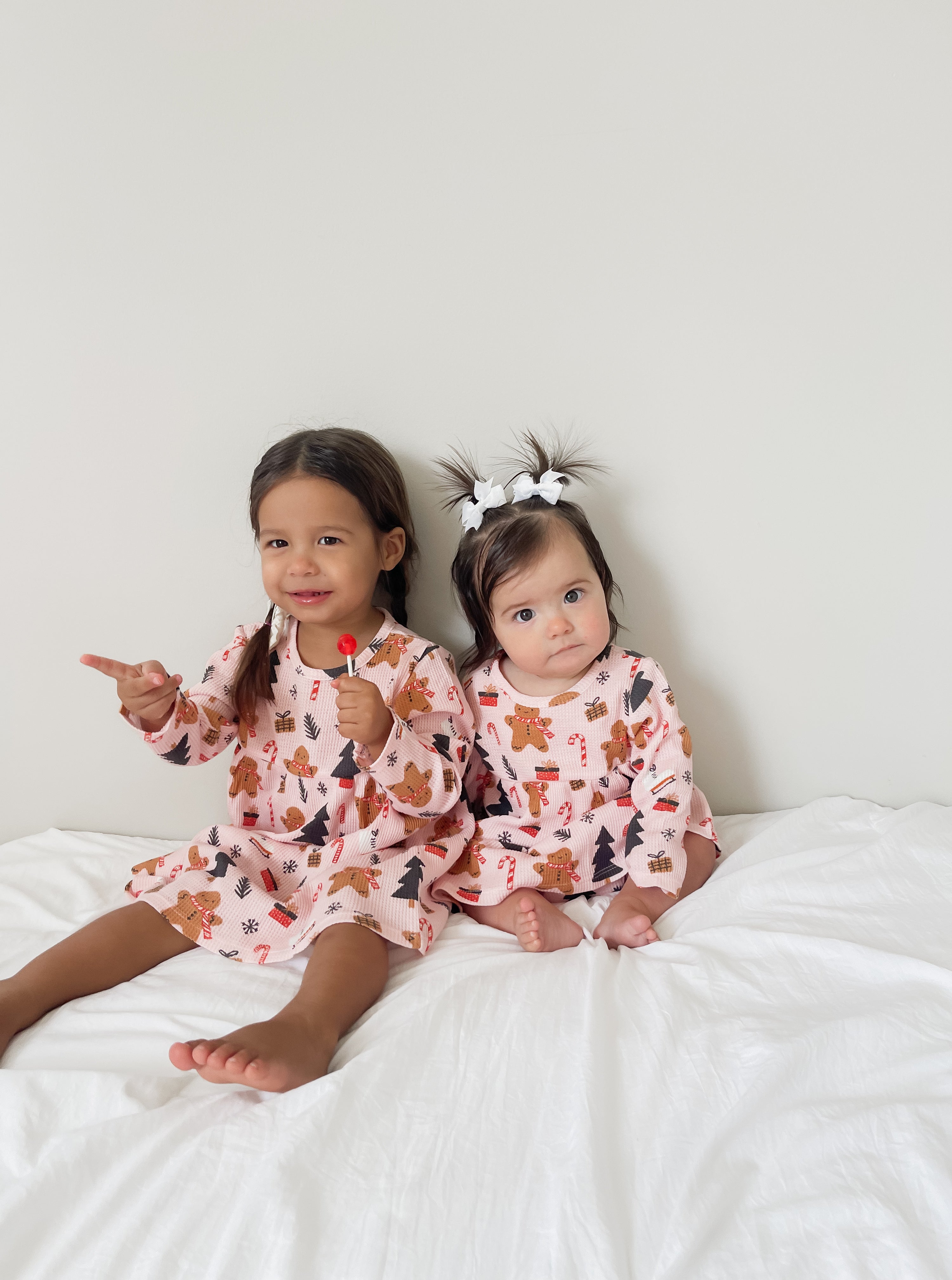 Two toddlers in festive pajamas sit on a white bed, one holding a lollipop and smiling at the camera.