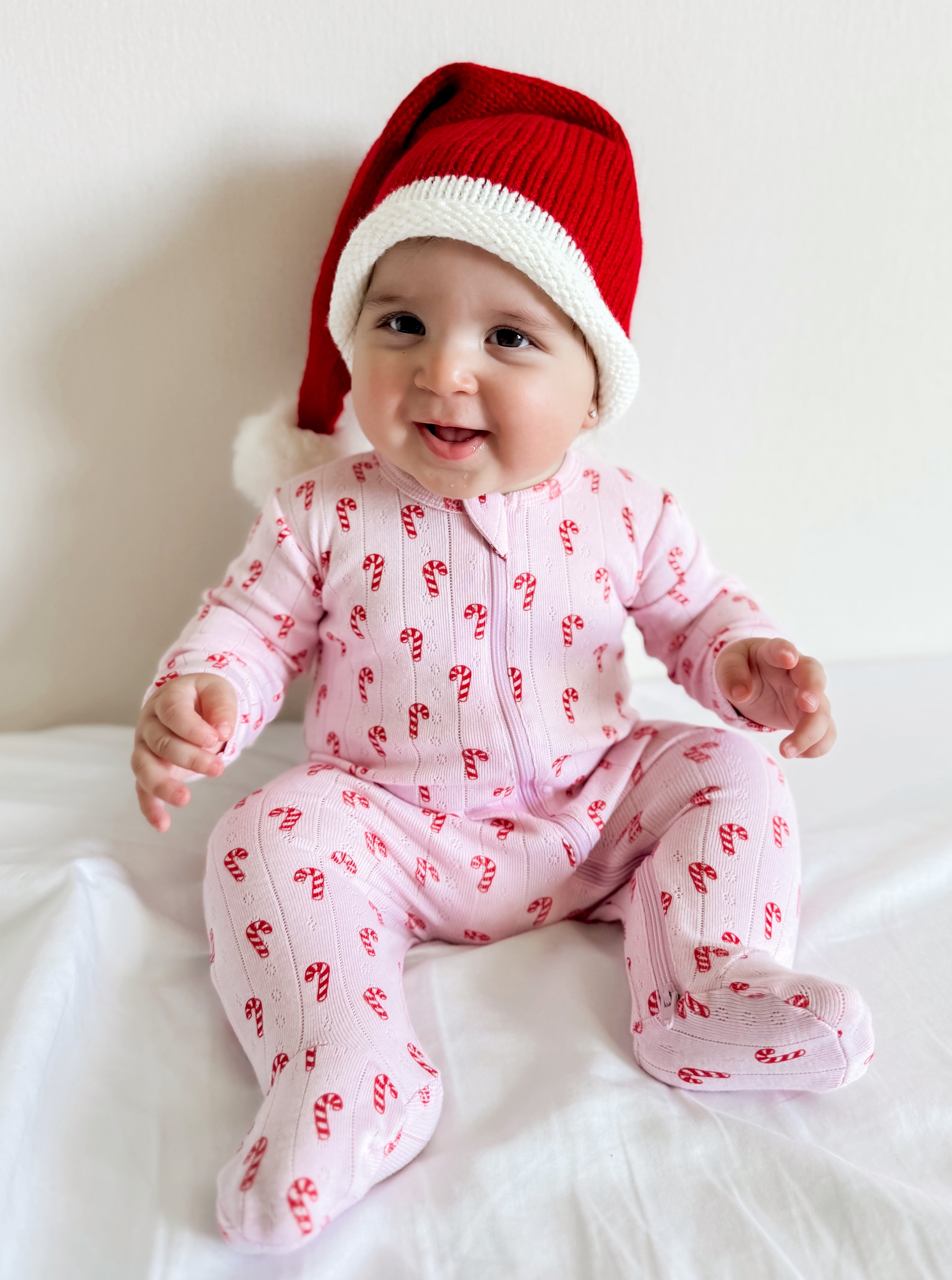 Smiling baby in a candy cane patterned onesie and red Santa hat, sitting on a white background.
