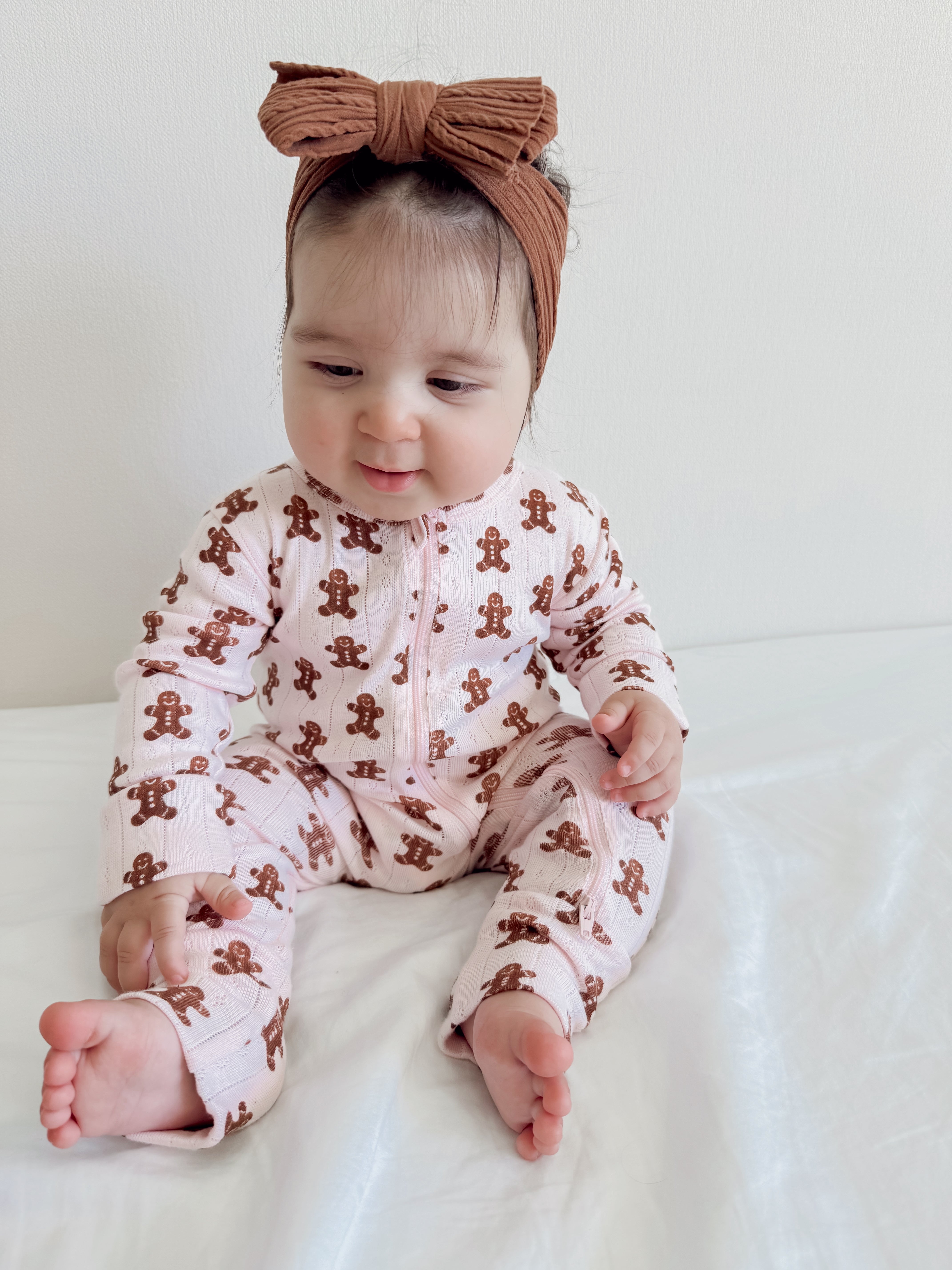 Baby in a pink gingerbread-patterned onesie and brown bow headband, sitting on a white surface.