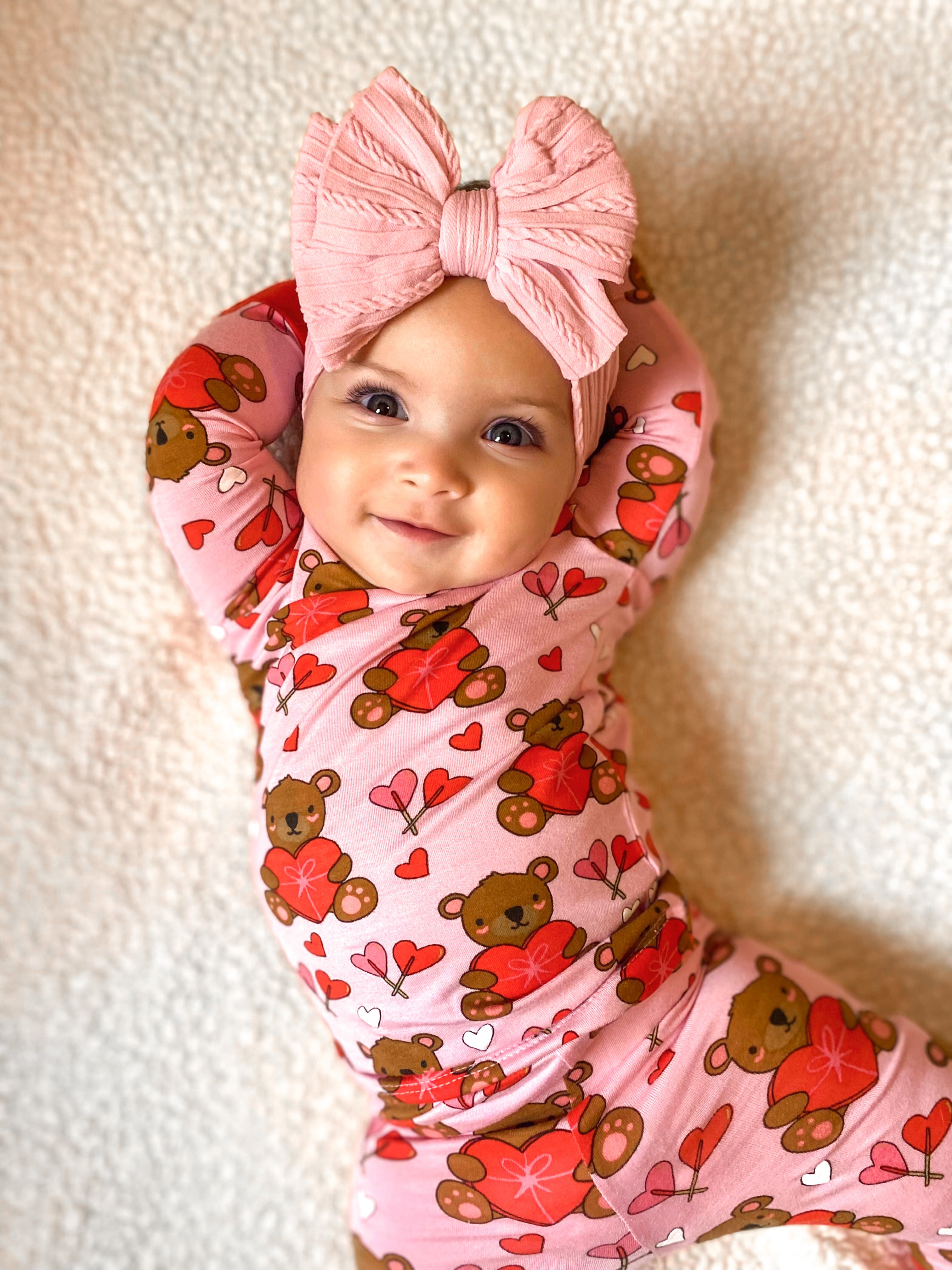 Smiling baby in pink bear-patterned outfit and large pink bow, lying on a soft white blanket.