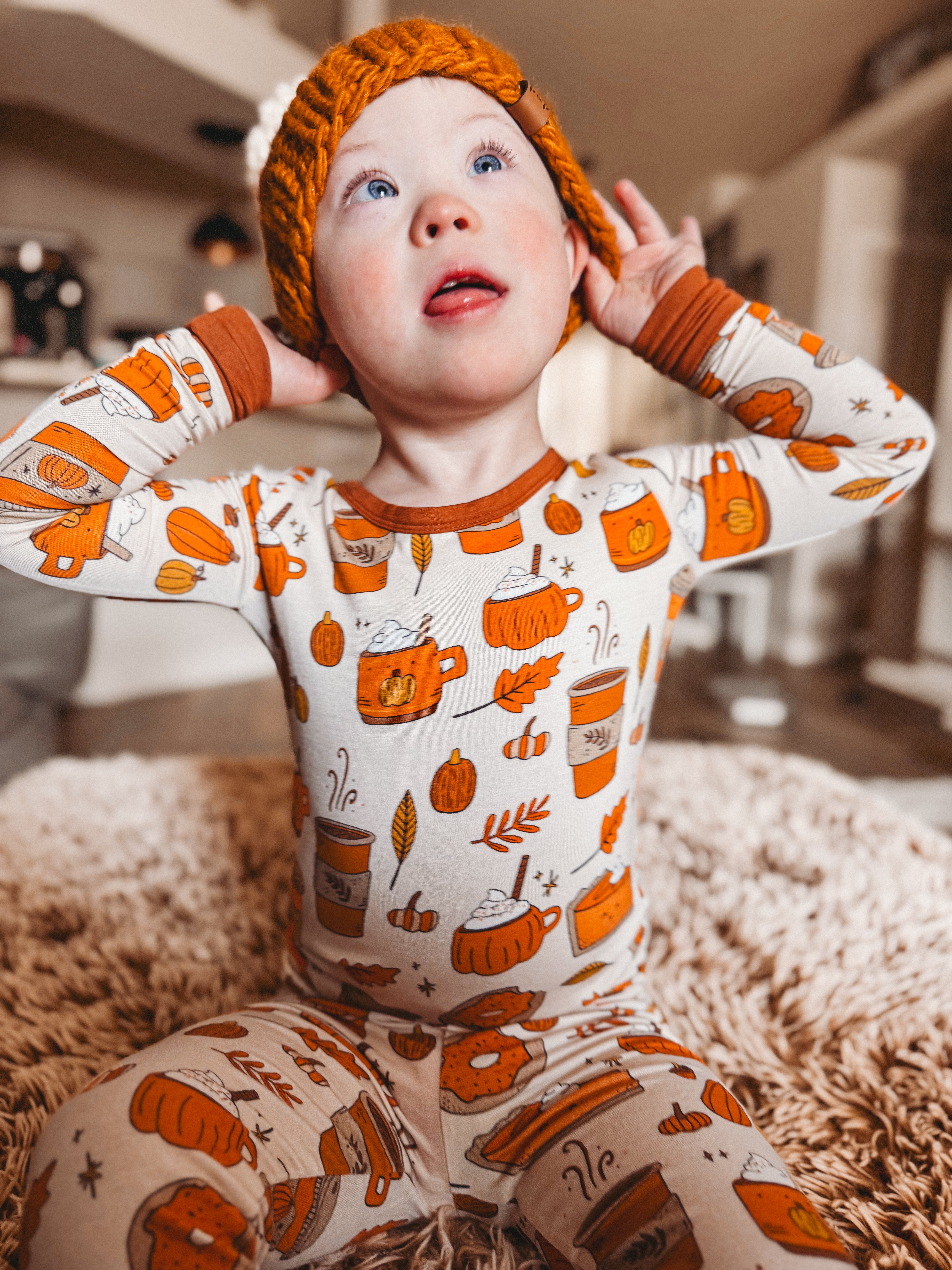 Young child in cozy autumn pajamas with pumpkin and beverage designs, wearing an orange knit hat, looking excited.