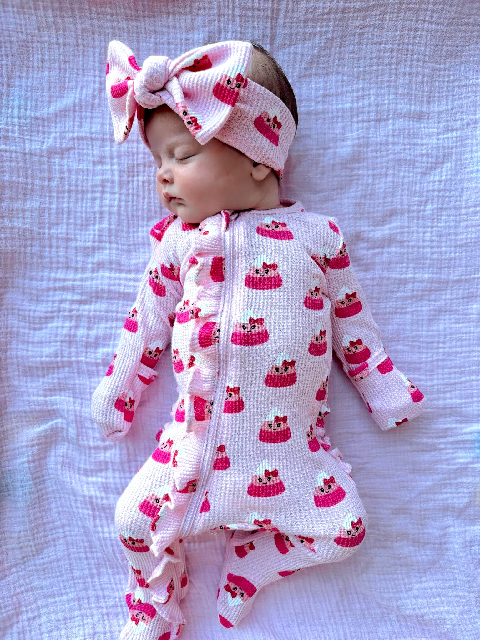 Infant wearing pink dessert-patterned pajamas and matching headband, sleeping on a white textured blanket.