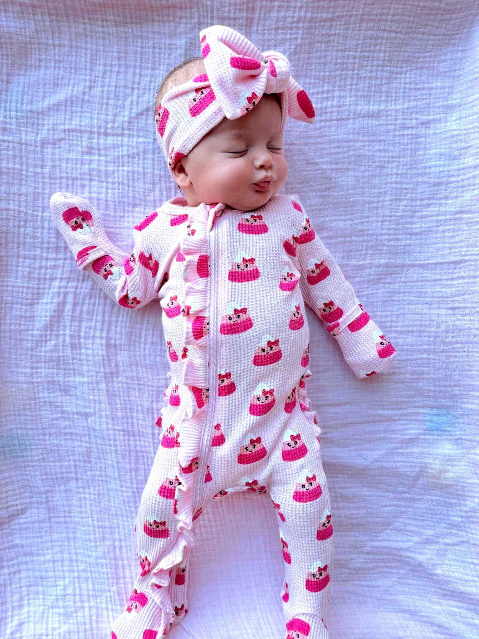 Baby wearing a pink cake-patterned onesie with a matching headband, lying on a white blanket.