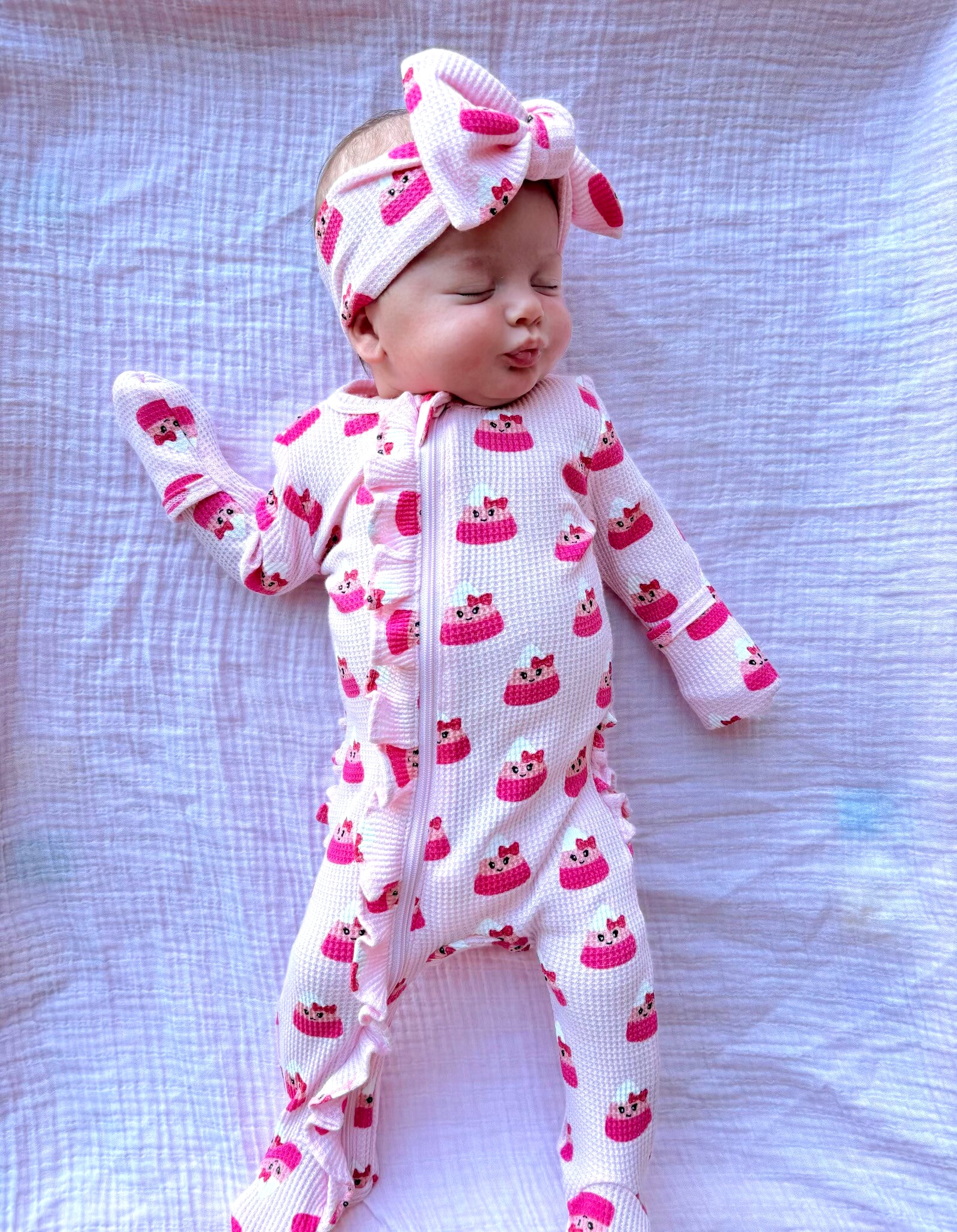 Baby wearing a pink cake-patterned onesie with a matching headband, lying on a white blanket.