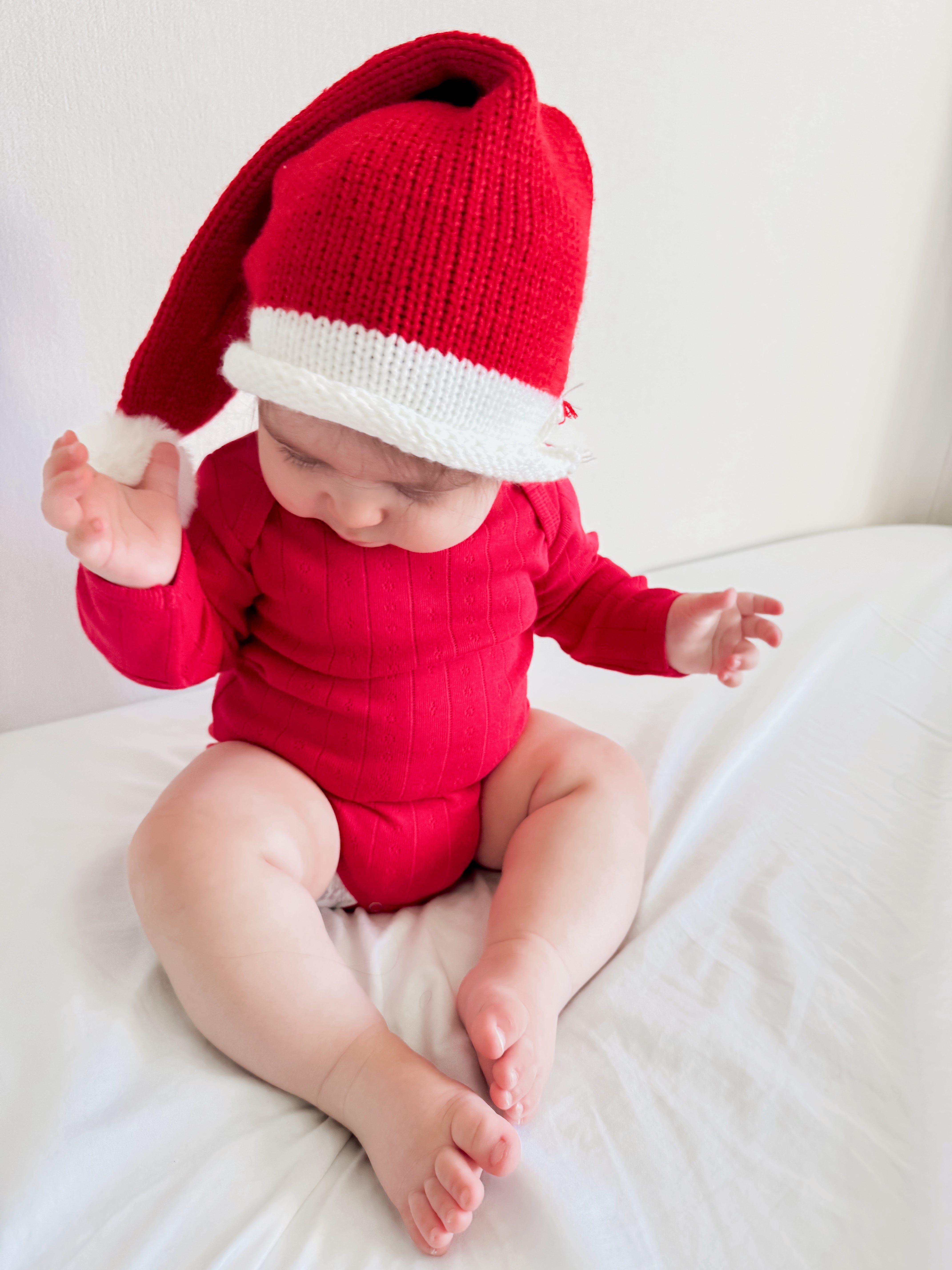 Baby in a red outfit and festive hat sitting on a bed, playing with hands and smiling.