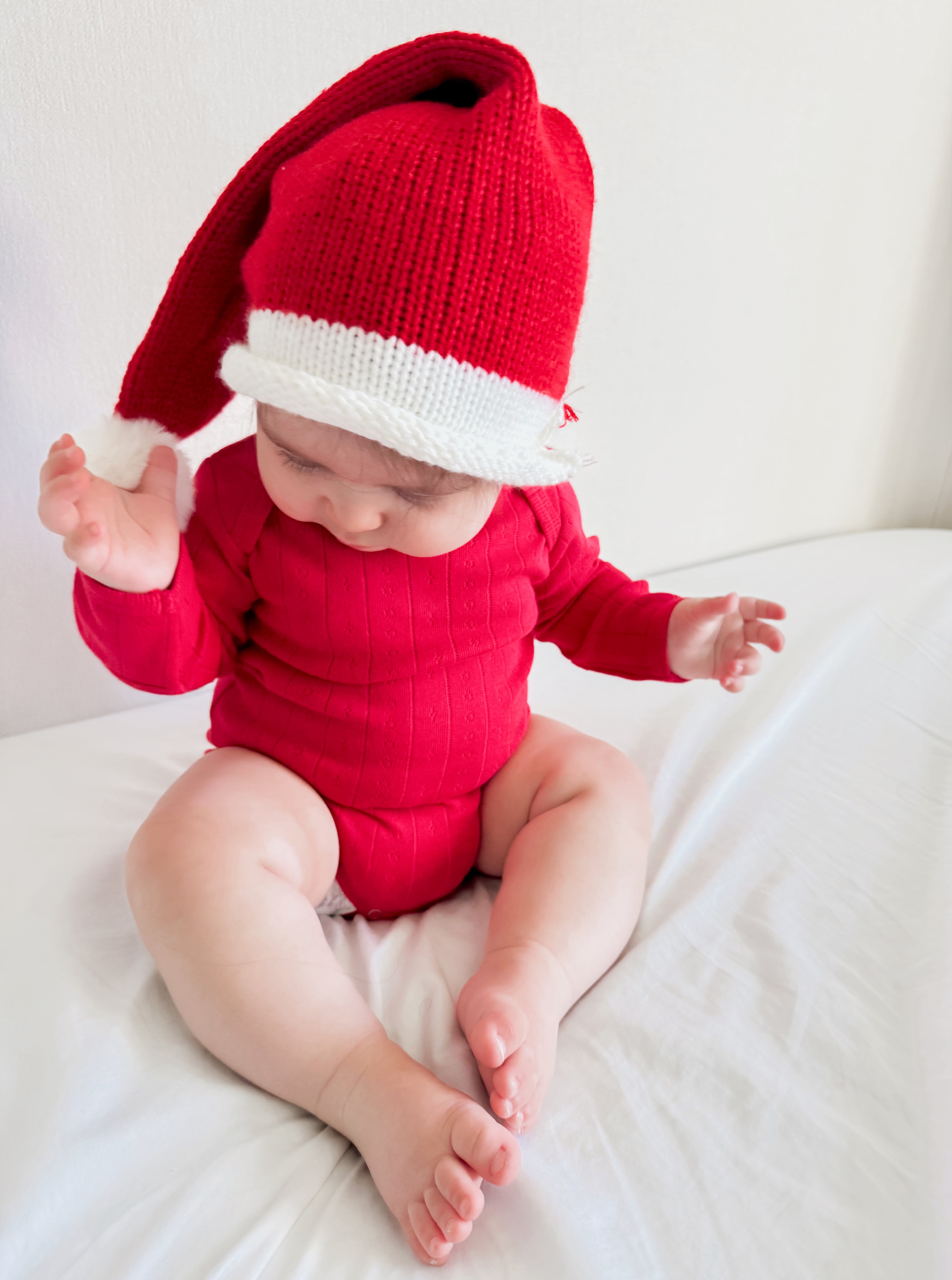 Baby in a red outfit and festive hat sitting on a bed, playing with hands and smiling.