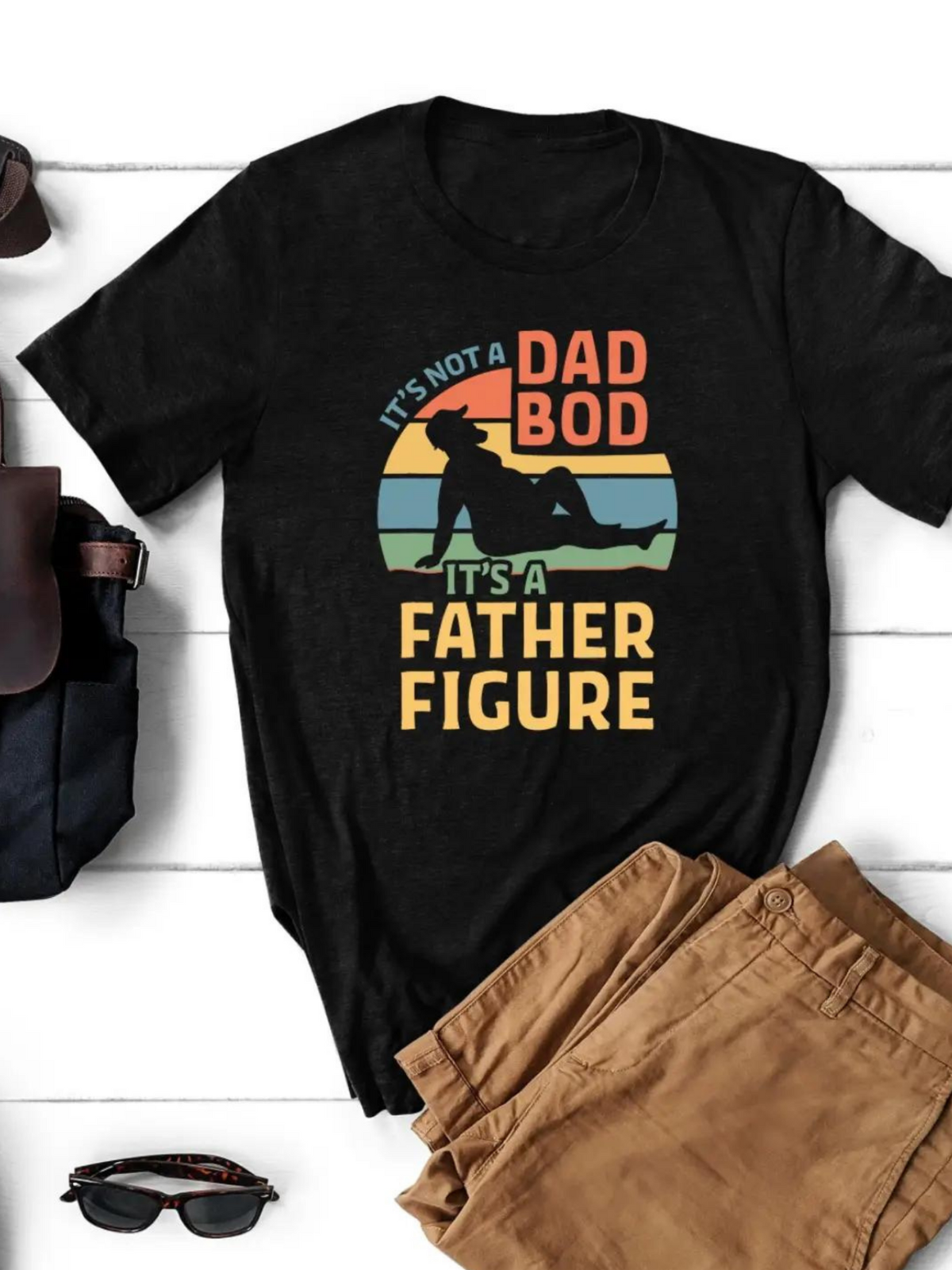 Black t-shirt with colorful text: "It's not a Dad Bod, it's a Father Figure." Laid on a wooden surface with sunglasses and pants.