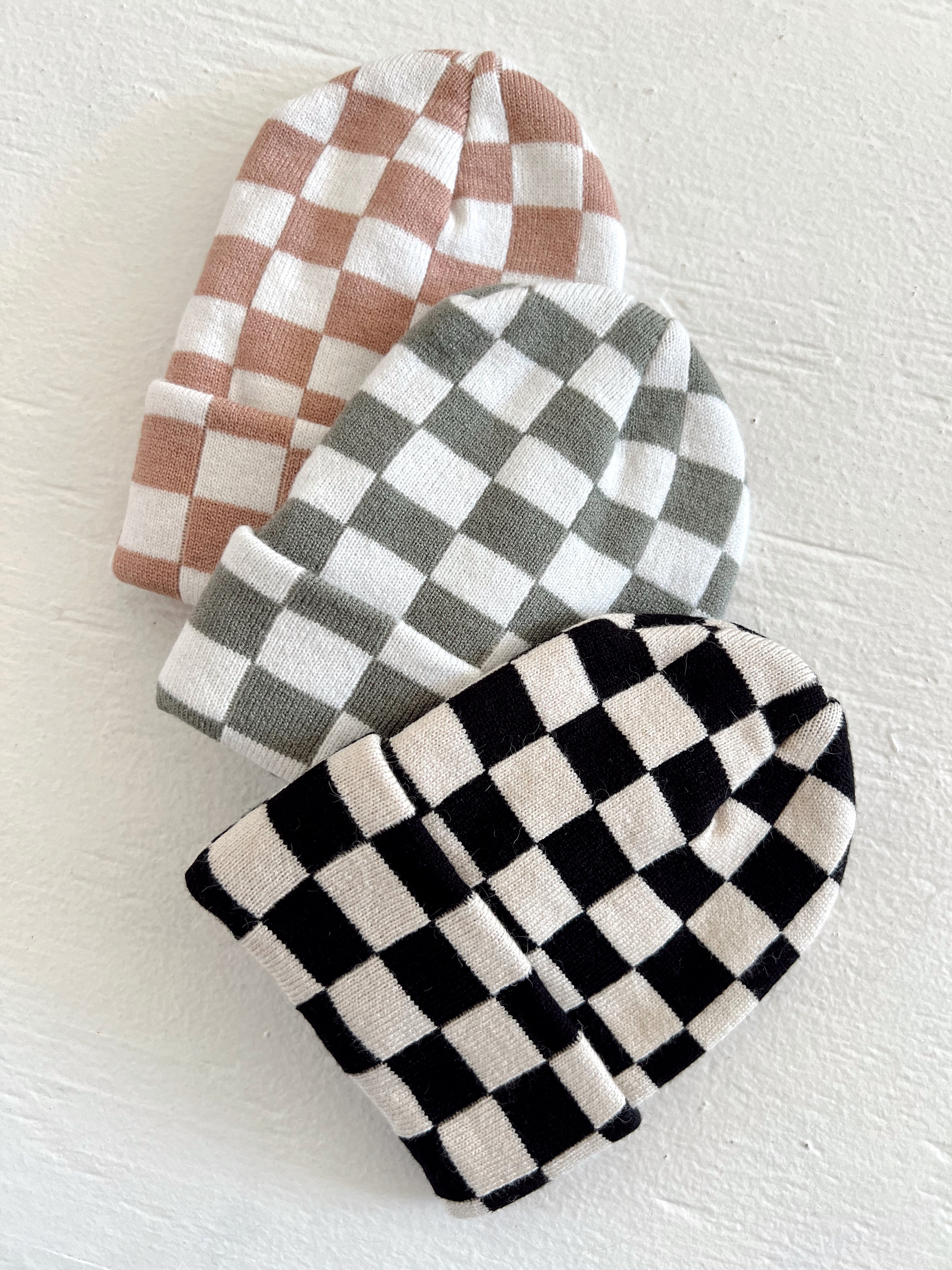 Three beanie hats in checkered patterns: blush pink, gray, and black with white, stacked on a white background.