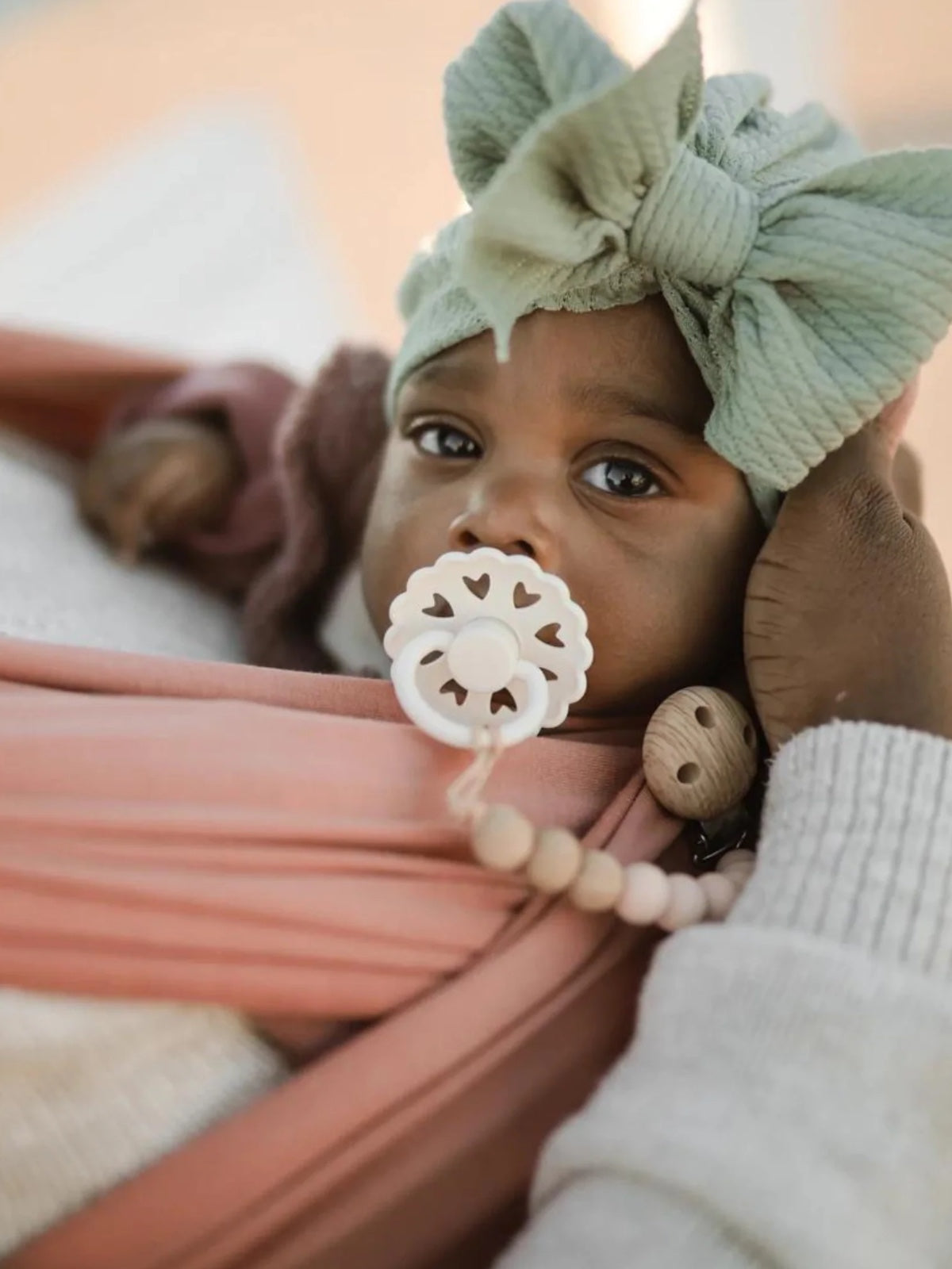 Infant in a mint bow headband holds a pacifier, wrapped in a soft carrier, with a gentle hand nearby.