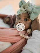 Infant in a mint bow headband holds a pacifier, wrapped in a soft carrier, with a gentle hand nearby.