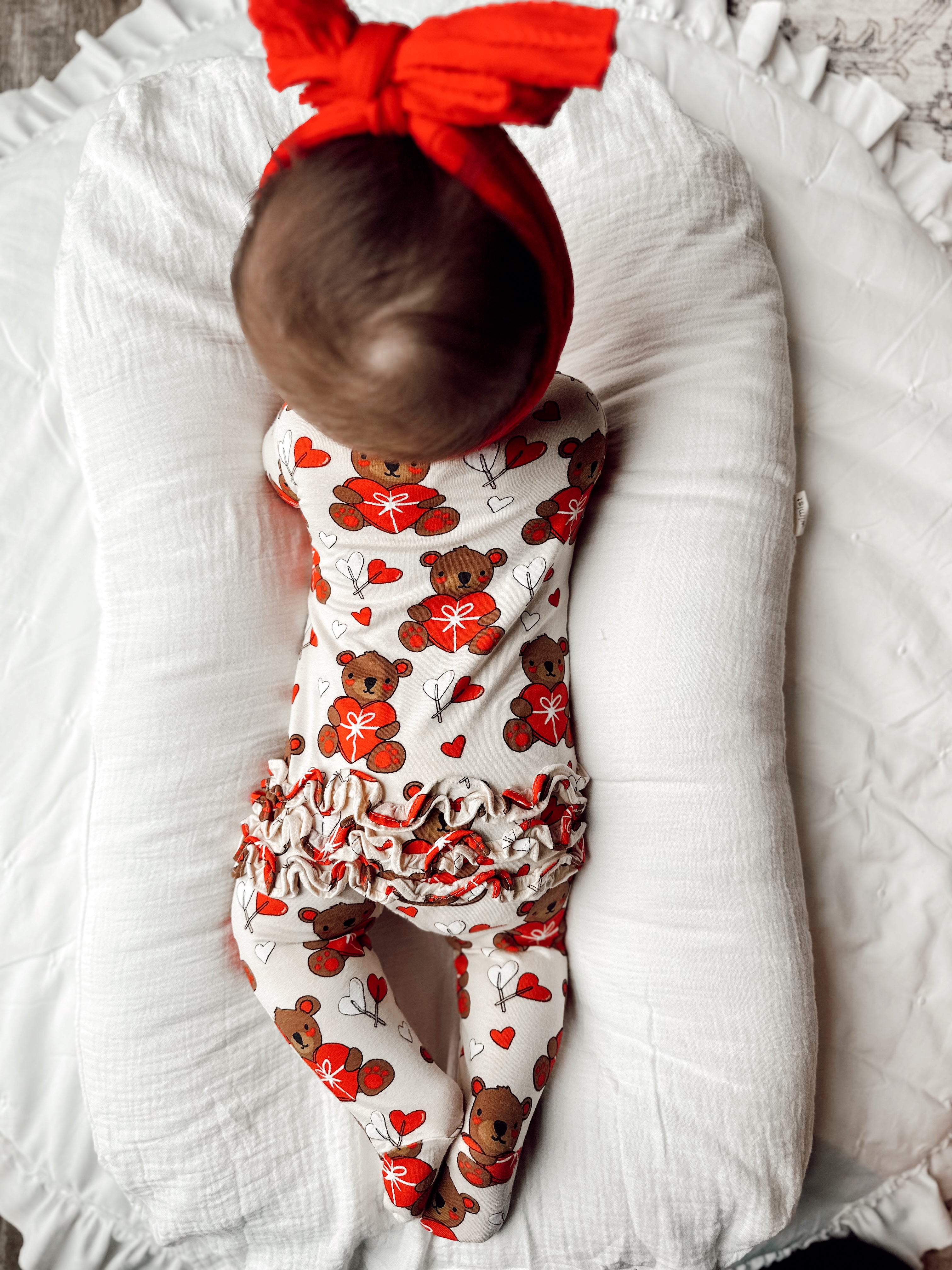 Baby lying on a white pillow, wearing a teddy bear-patterned outfit and a red headband.