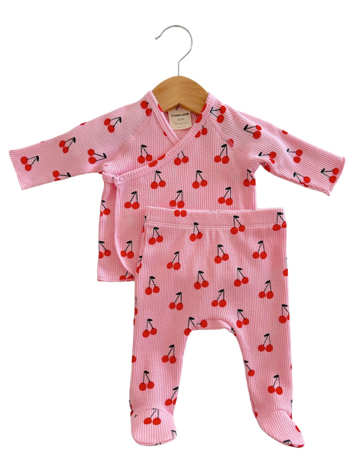 Pink baby outfit with cherry pattern, featuring a long-sleeve top and matching pants, hanging on a wooden hanger.