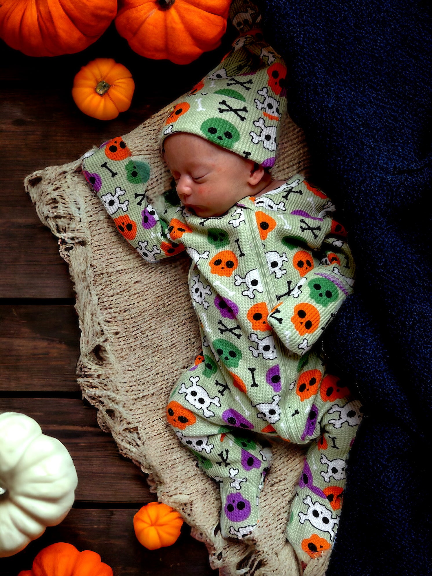 Sleeping baby in a colorful skeleton outfit surrounded by pumpkins and a cozy blanket.
