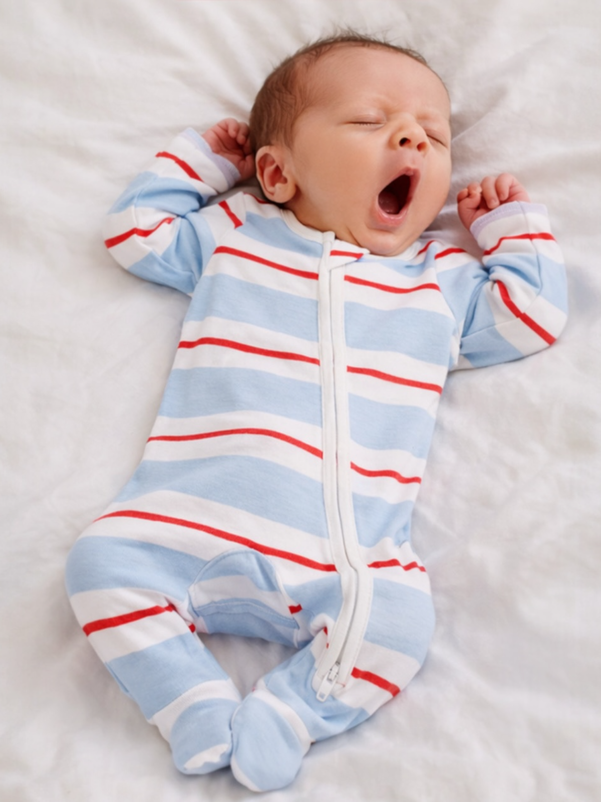 Sleeping newborn yawning in a striped blue and white onesie on a soft, white blanket.