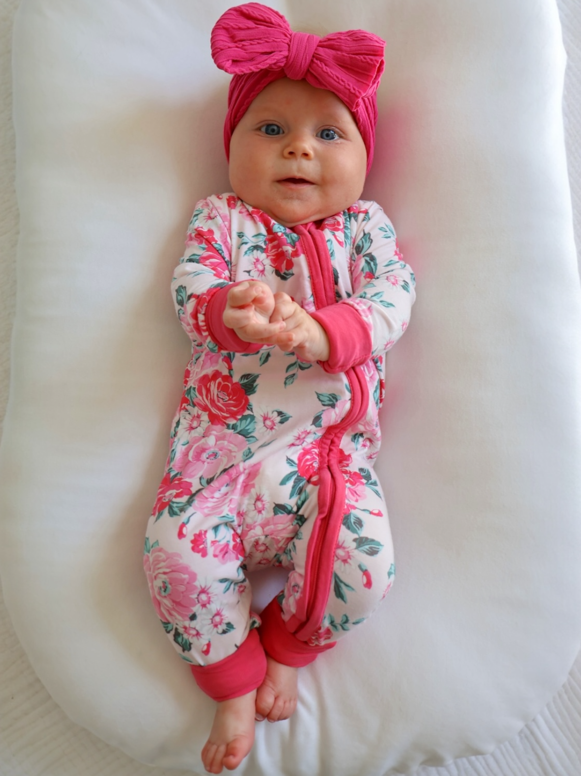 Infant in floral onesie and pink headband, lying on a white cushion, smiling and playing with hands.