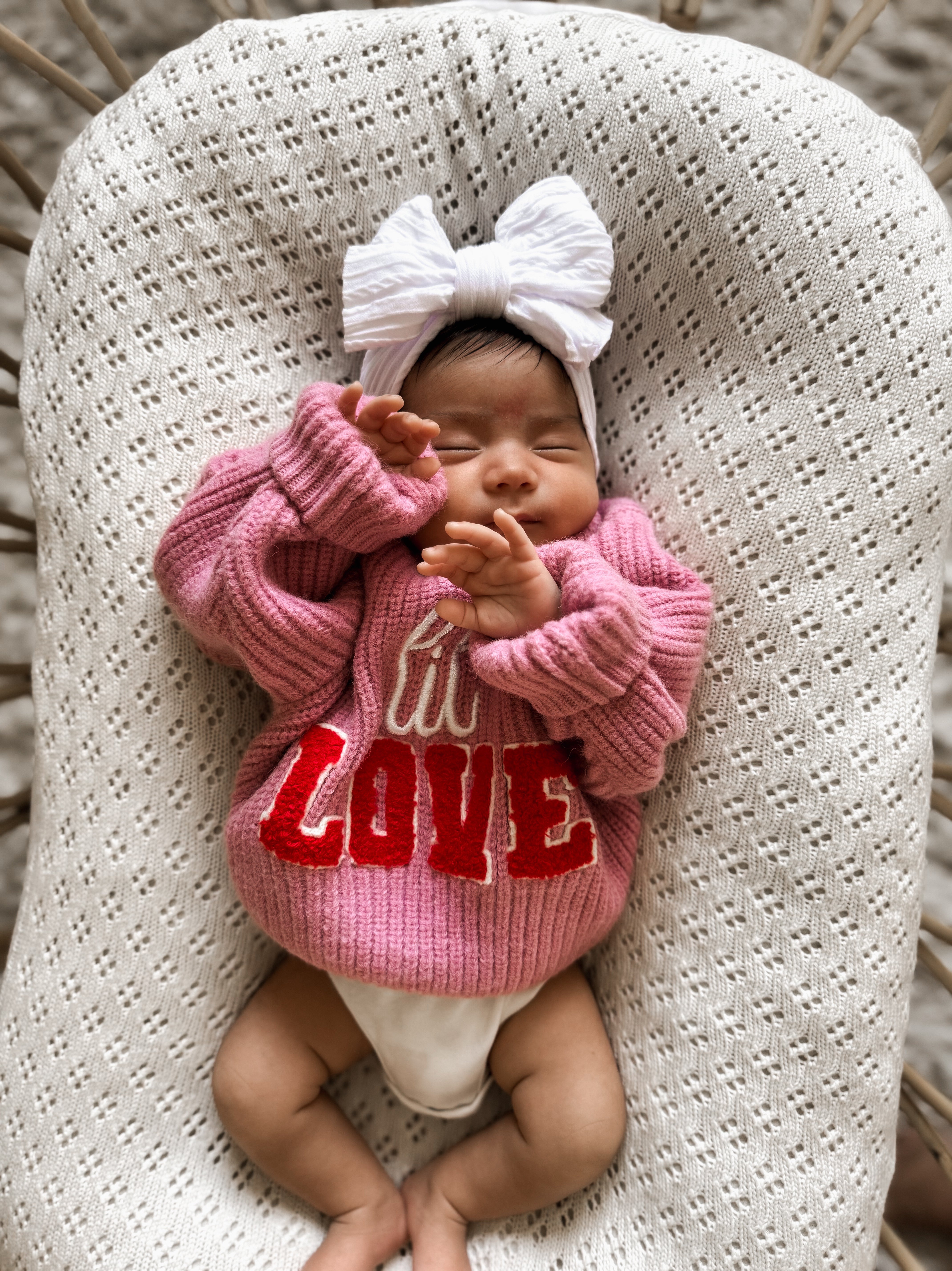 Newborn baby in a pink sweater with red "LOVE" lettering, wearing a white bow, lying on a textured blanket.