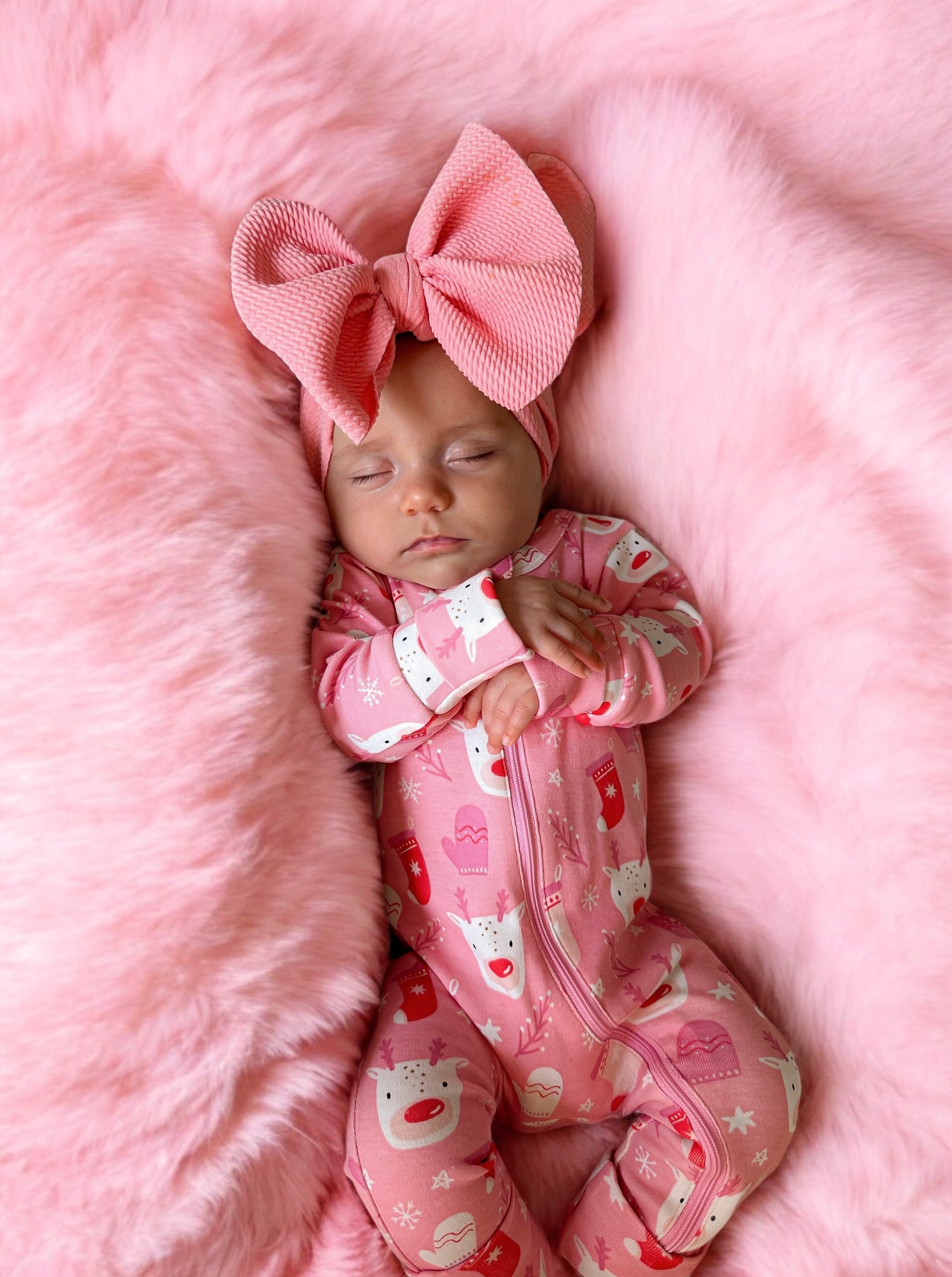 Sleeping baby in pink pajamas with a large pink bow on a fluffy pink blanket.