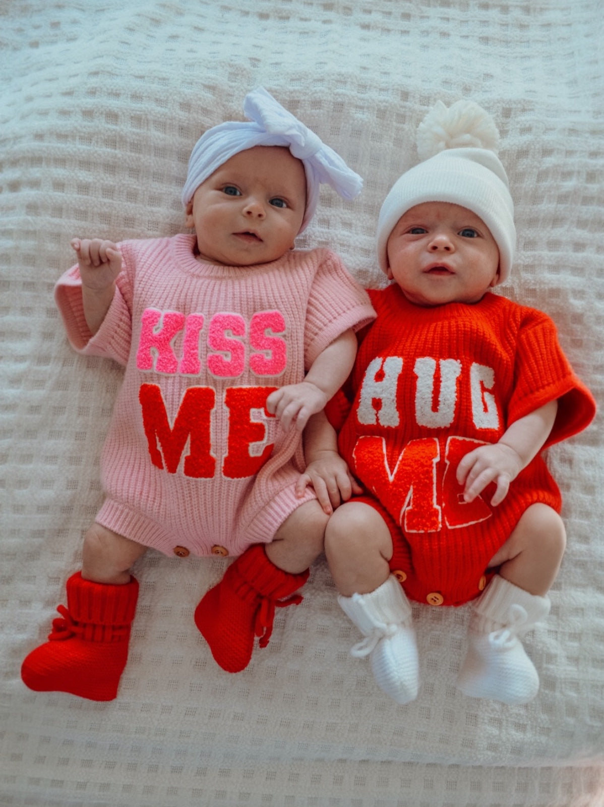 Two babies in colorful outfits that say "KISS ME" and "HUG ME," sitting together on a soft blanket.
