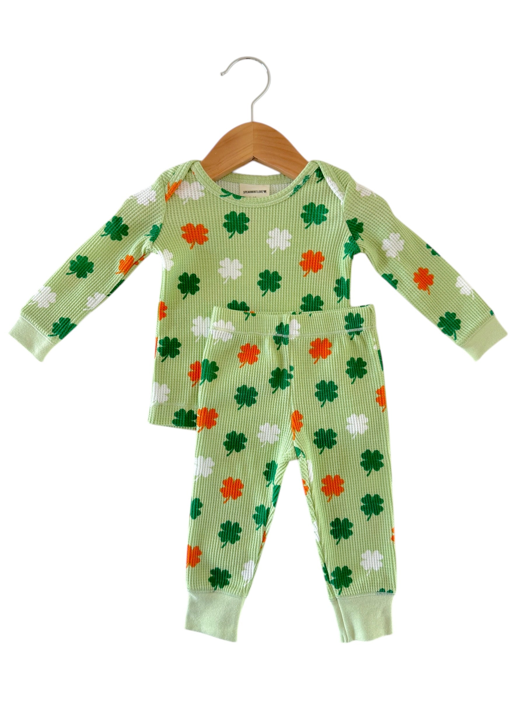 Green children's pajamas with shamrock patterns in green, white, and orange, hanging on a wooden hanger.
