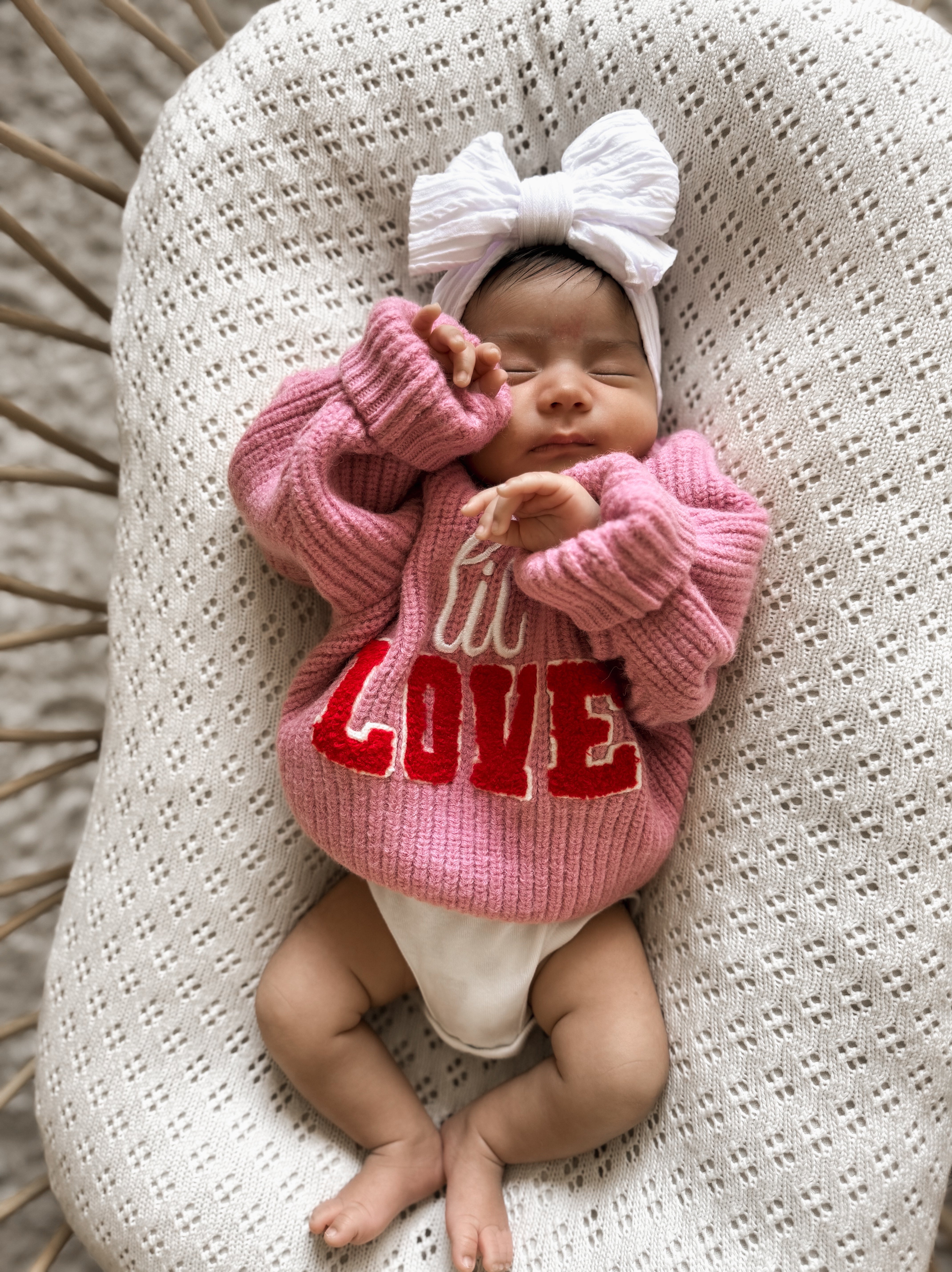 Baby girl in a pink sweater with "LOVE" text, wearing a large white bow, resting peacefully on a textured blanket.