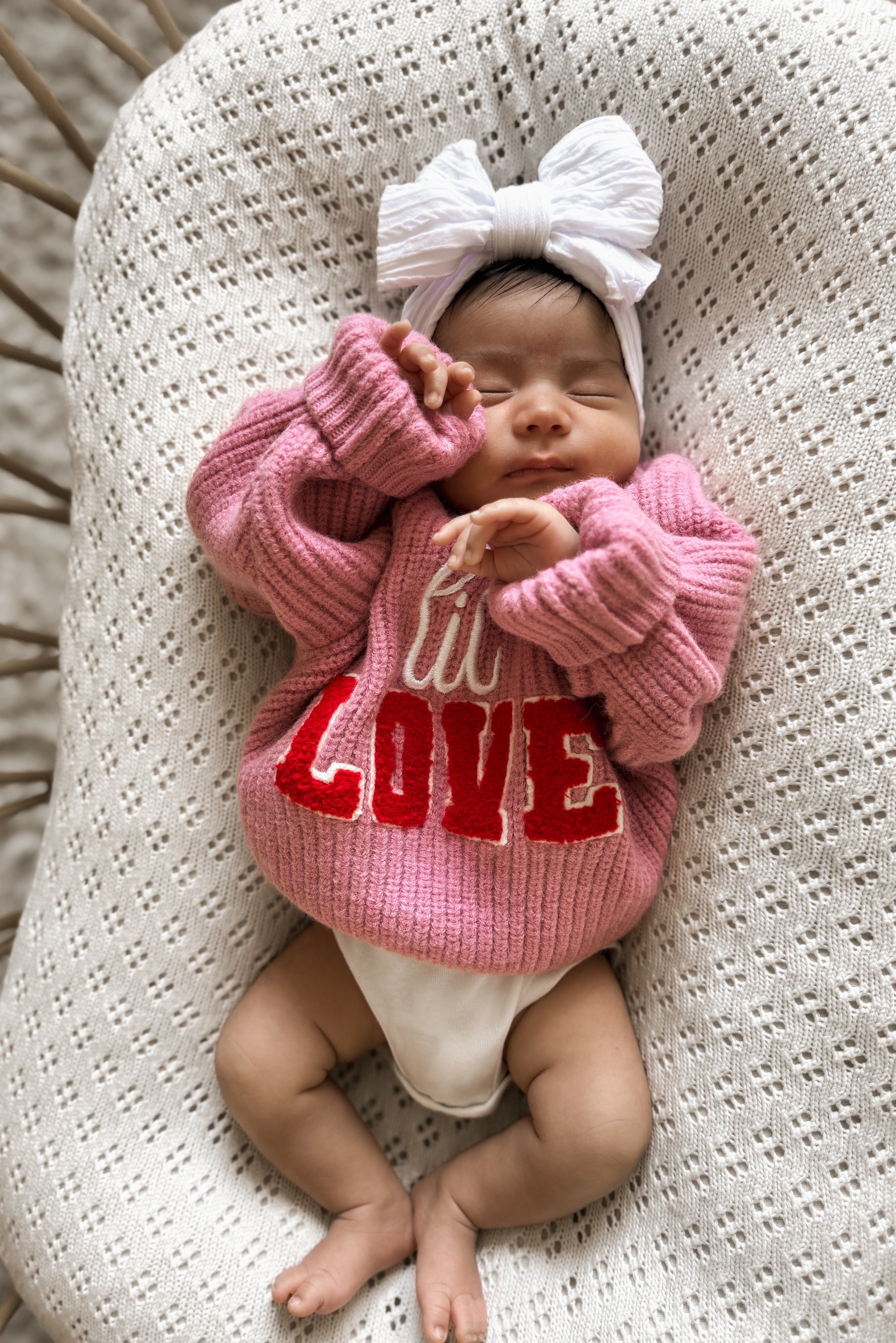 Baby girl in a pink sweater with "LOVE" text, wearing a large white bow, resting peacefully on a textured blanket.