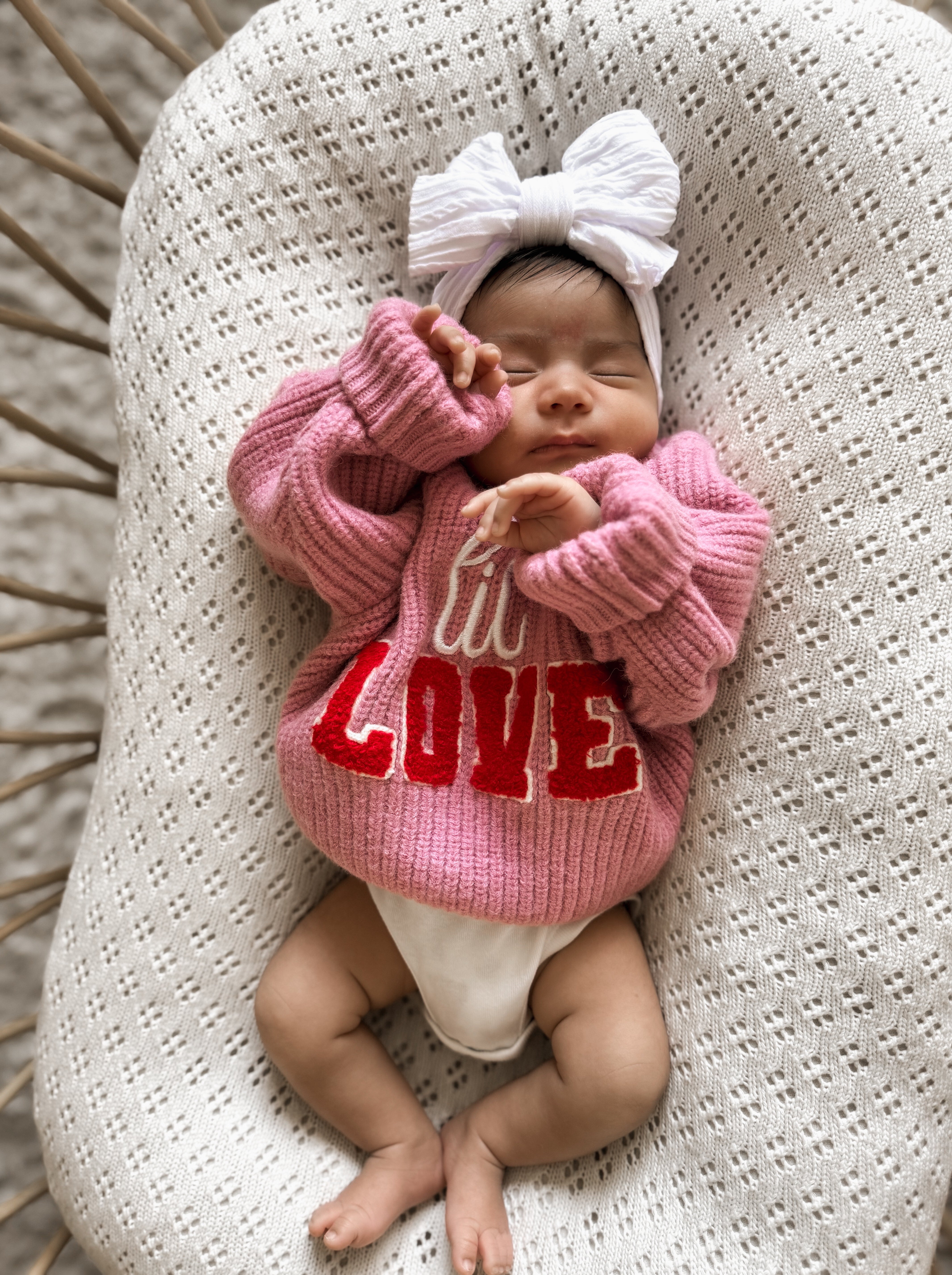 Baby girl in a pink sweater with "LOVE" text, wearing a large white bow, resting peacefully on a textured blanket.