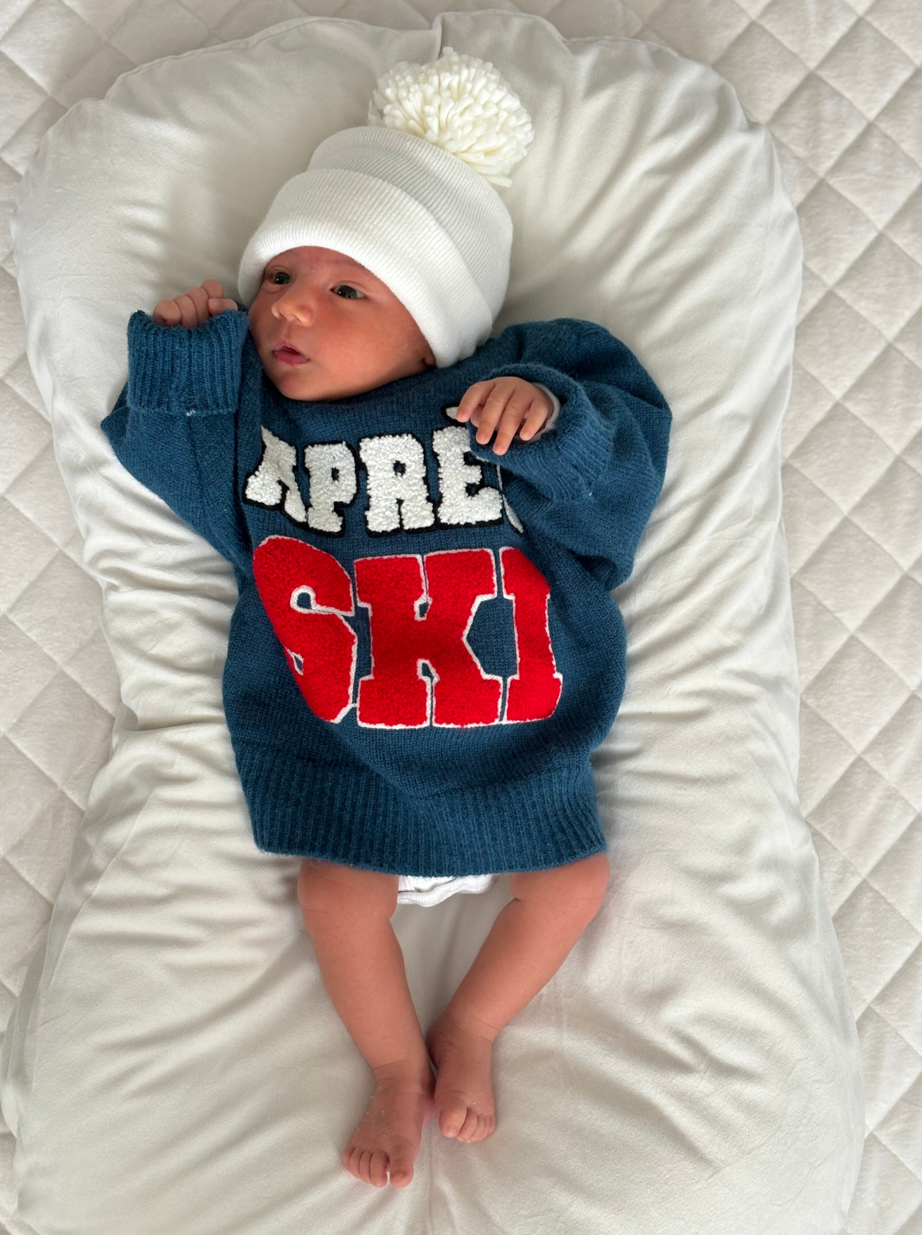 Infant in a blue sweater with "Apres Ski" text and a white pom-pom hat, lying on a quilted blanket.